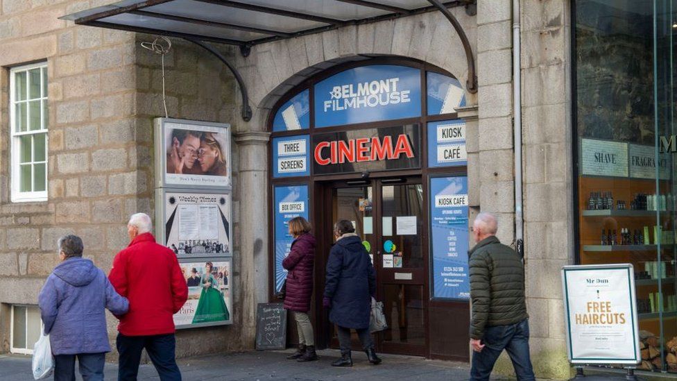 The outside of the Belmont Filmhouse, with five people walking past the door in two pairs and an individual. There are film posters around the door and box office information.