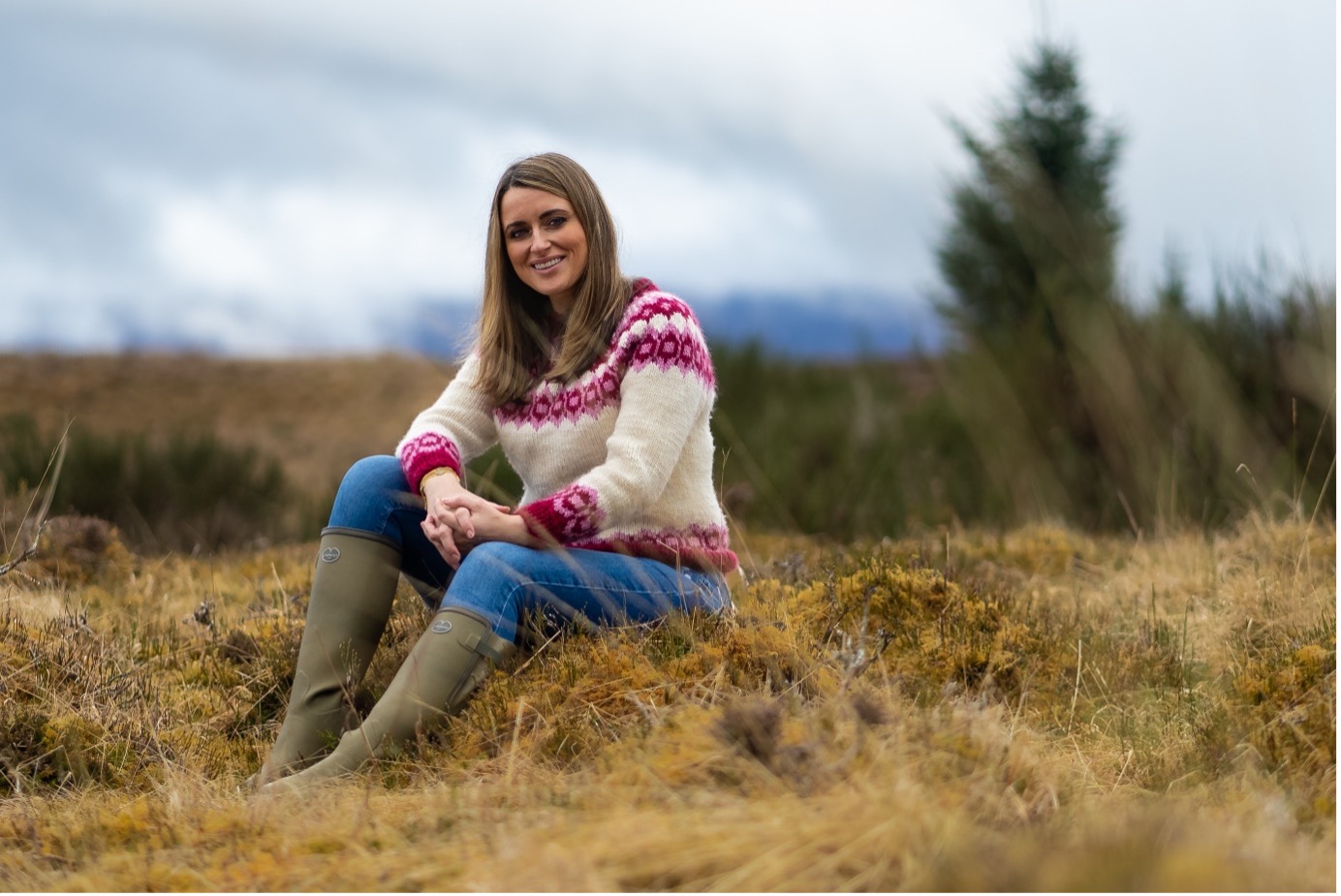 Promotional shot from Secrets in the Peat, courtesy of the BBC. Presenter Anne McAlpine wears wellington boots, jeans and a white wooden jumper, and sits on grass in an outdoor location.