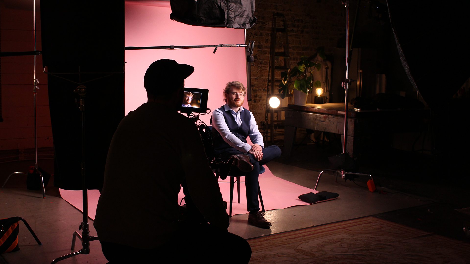 Still of Jonathan Mullen in Working Differently, credit: The Portal Studios. Jonathan sits on a chair against a pink background, with a man holding a camera filming him, with his back to the camera.