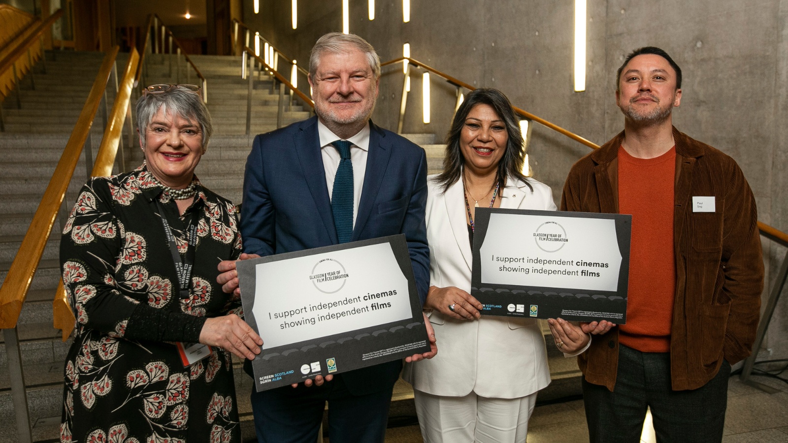 Credit Eoin Carey, courtesy GFF. The image shows four people standing in a row from left to right: CEO of Glasgow Film of Glasgow Film Allison Gardner, Cabinet Secretary for Constitution, External Affairs and Culture Angus Robertson MSP, Glasgow Kelvin MSP and Minister (designate) for Culture, Europe and International Development, Kaukab Stewart, and filmmaker Paul Sng. They are holding two signs which read I support independent cinema showing independent films