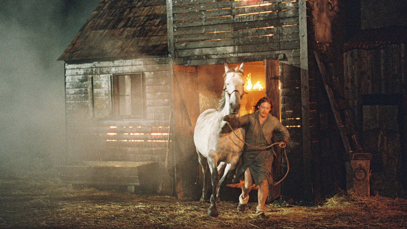 Still from Harvest, credit Jaclyn Martinez. Actor Caleb Landry-Jones leads a white horse by the reigns away from a burning wooden building at night