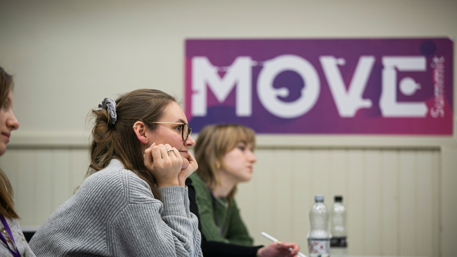 A woman wearing glasses and a light blue jumper sits facing to the right beside a sign that reads MOVE in capital letters