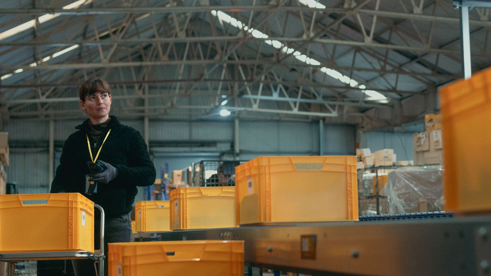 Still from On Falling, courtesy of Sixteen Films. A woman, played by actress Joana Santos stands in a large warehouse, with yellow crates stacked on tables all around her.