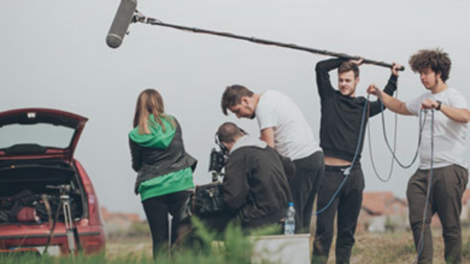 The image shows a film crew with equipment. From left there is the boot of a red car with equipment it in, a person wearing a black jacket and green hoodie with their back to the camera, a man siting holding a camera and another man also looking at the camera to his right. Then a man wearing black trousers and jumper is holding a mic in the air and is next to another man holding the cables of this, wearing a white t-shirt and brown trousers. They are outdoors with grass in the foreground of the image and houses in the background