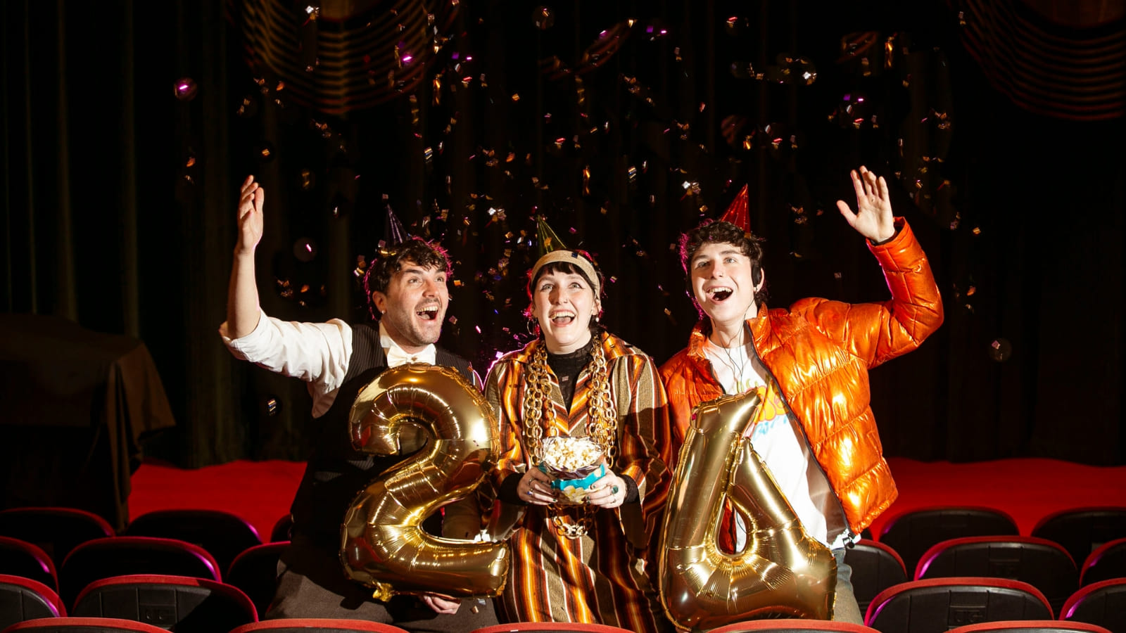Three people stand in a cinema screen, surrounded by chairs. They look exited, are smiling, and are holding a large gold balloon numbers 2 and 4. They have party hats on and there is glittery confetti falling from above.