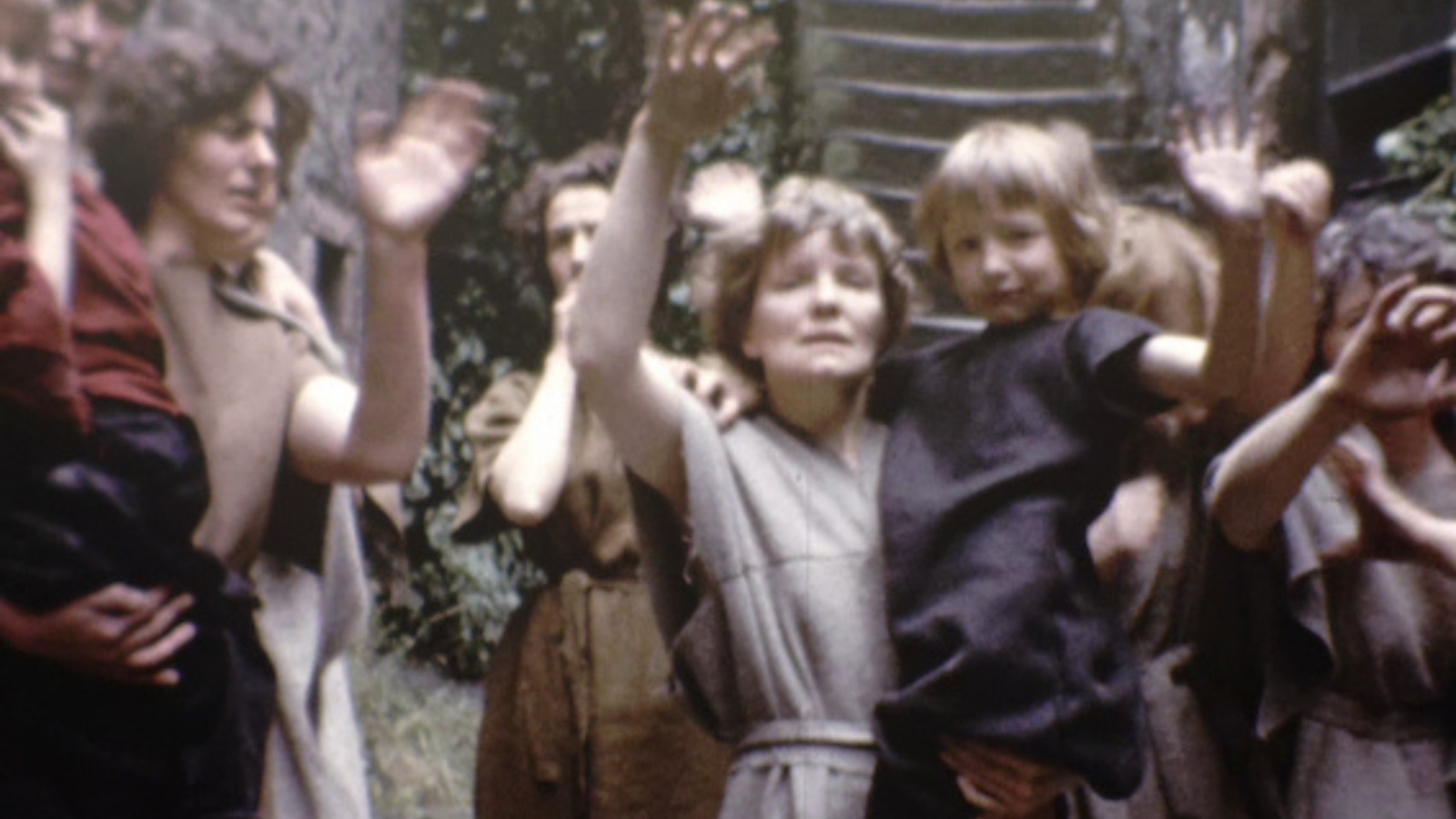Still from Sons of Heroes. A woman holding a small child in a crowd of people waves to someone off camera