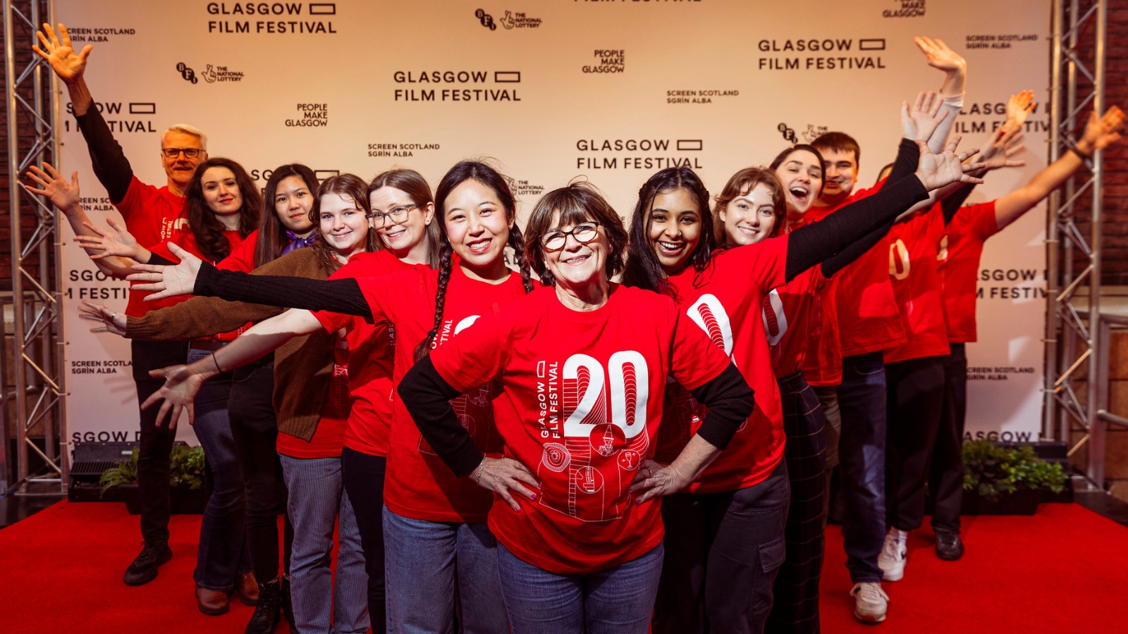 Volunteers wearing red t-shirts which read '20' on them stand in a v shape in front smiling at the camera