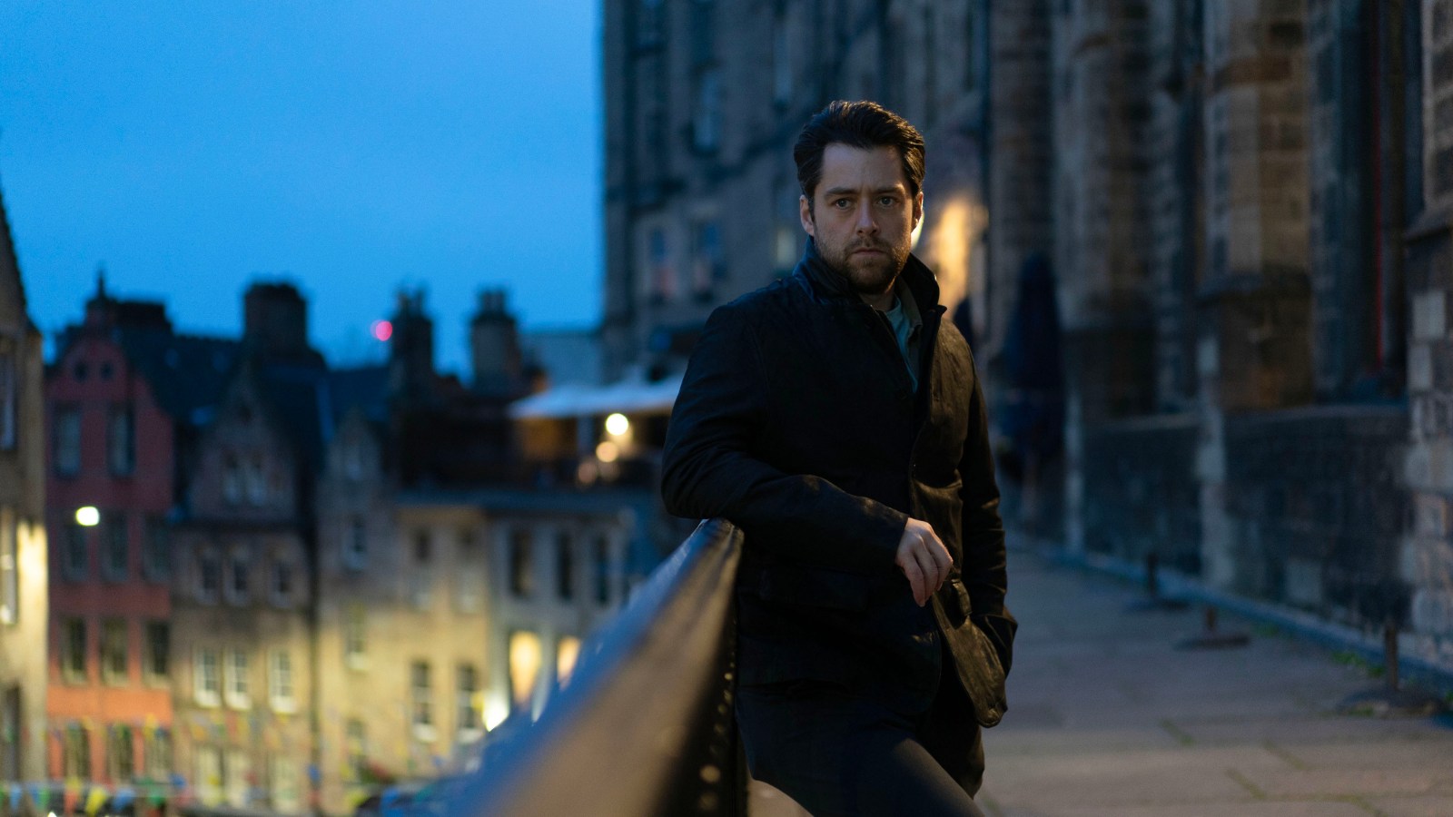 Still from Rebus, courtesy of BBC. Richard Rankin wears black jacket and trousers and leans on a railing on Victoria Terrace in Edinburgh.