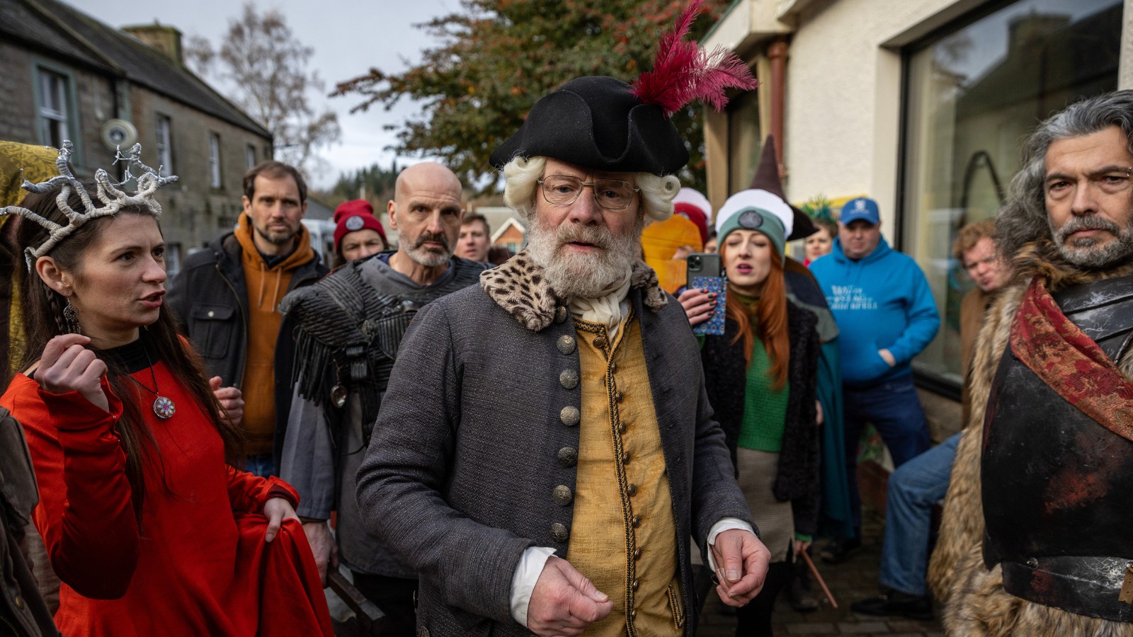 Still from The Fall of Douglas Weatherford, credit Saskia Coulson, which shows Kenneth (Peter Mullan) standing infant of a crowd of people. They all wear 18th century costumes.