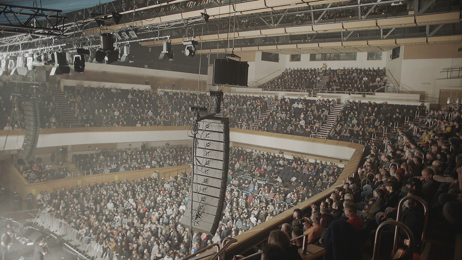 A large audience in a concert auditorium. Huge speakers hang from the roof.