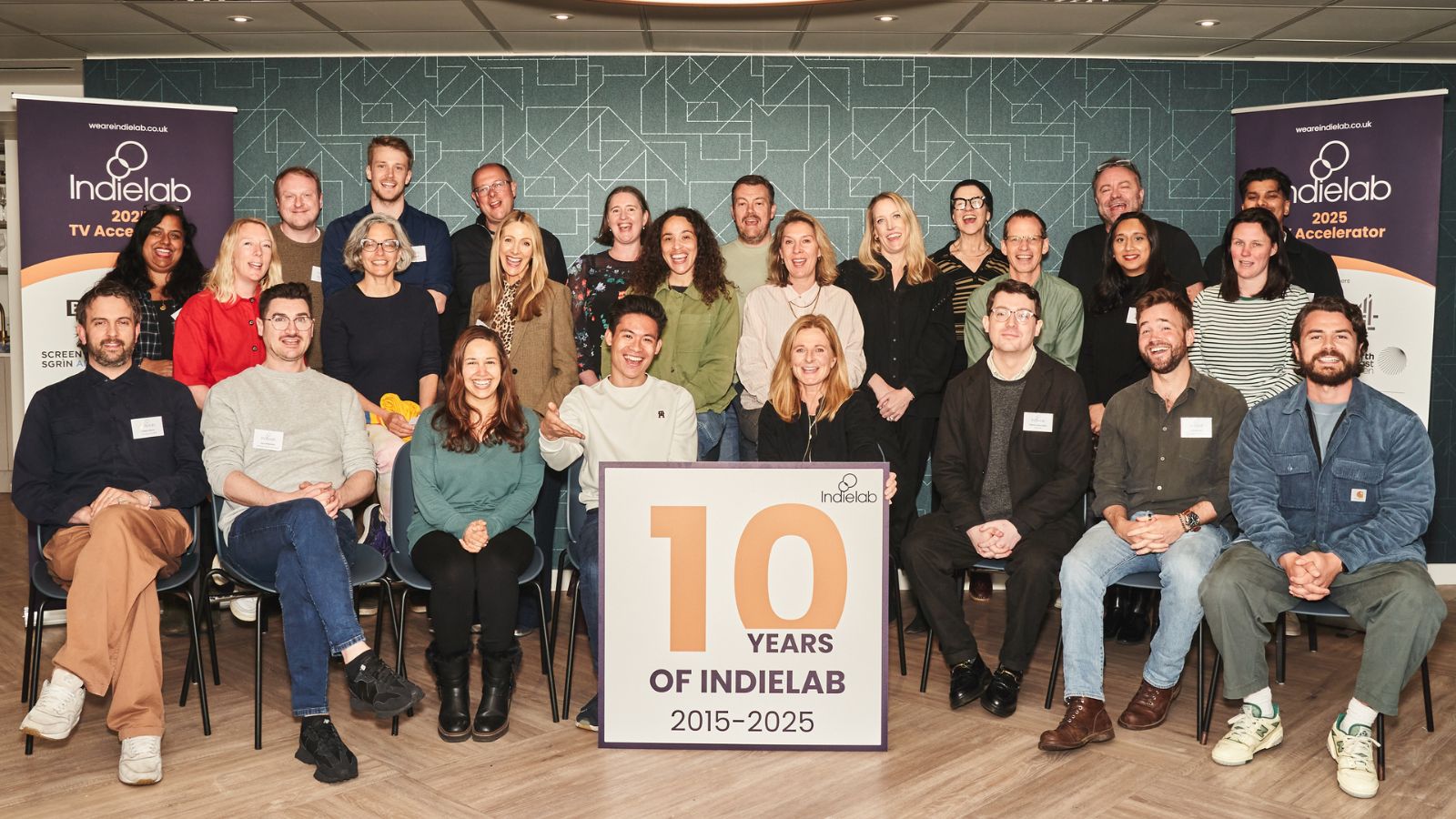 A group of people sit and stand in three rows indoors.