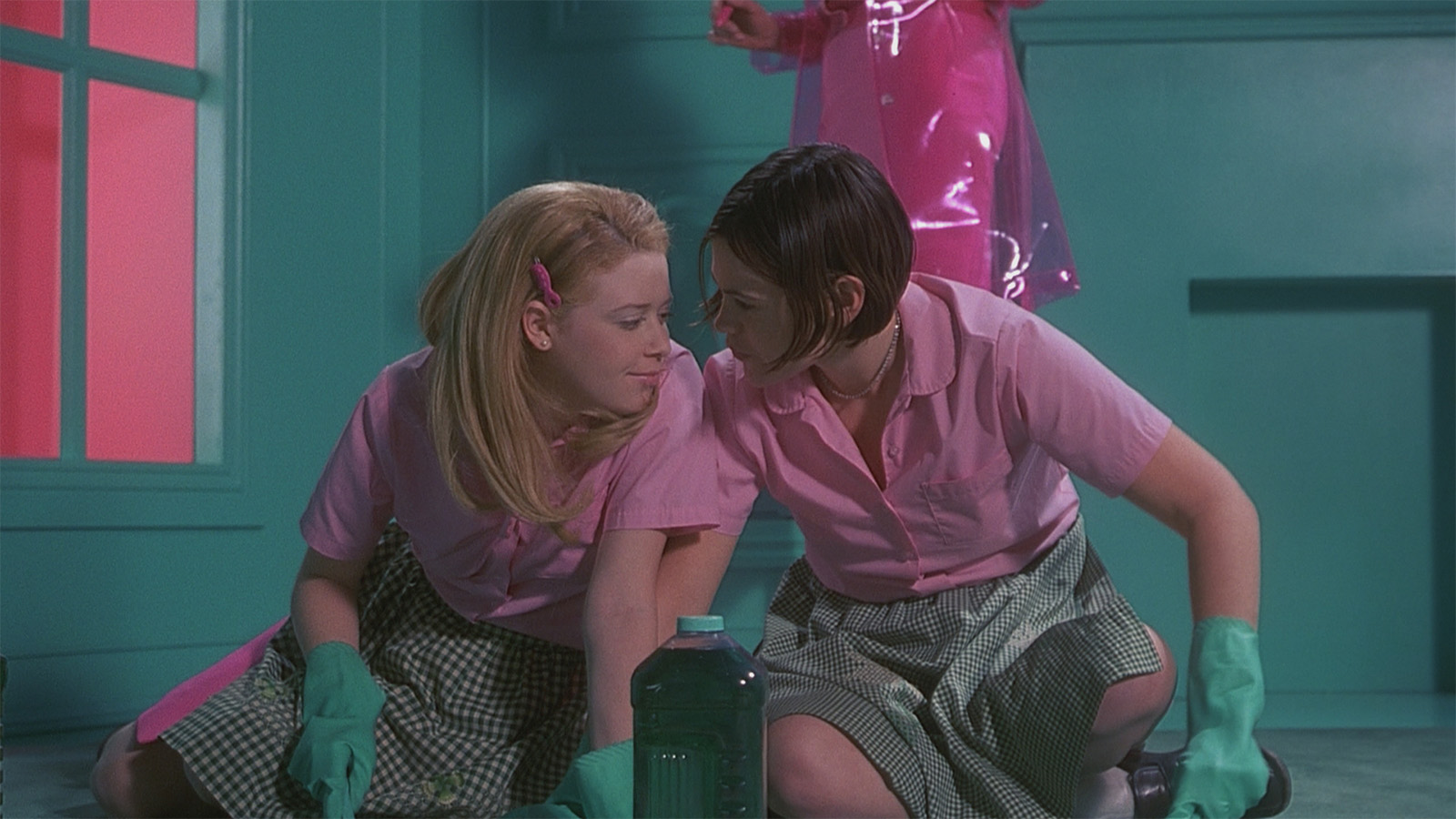 Two people wearing pink shirts, plaid skirts, and green gloves, lean close to each other while kneeling on the floor. A green cleaning bottle is in front of them. The background features teal walls and a pink window. This is a still from the film But I'm A Cheerleader.