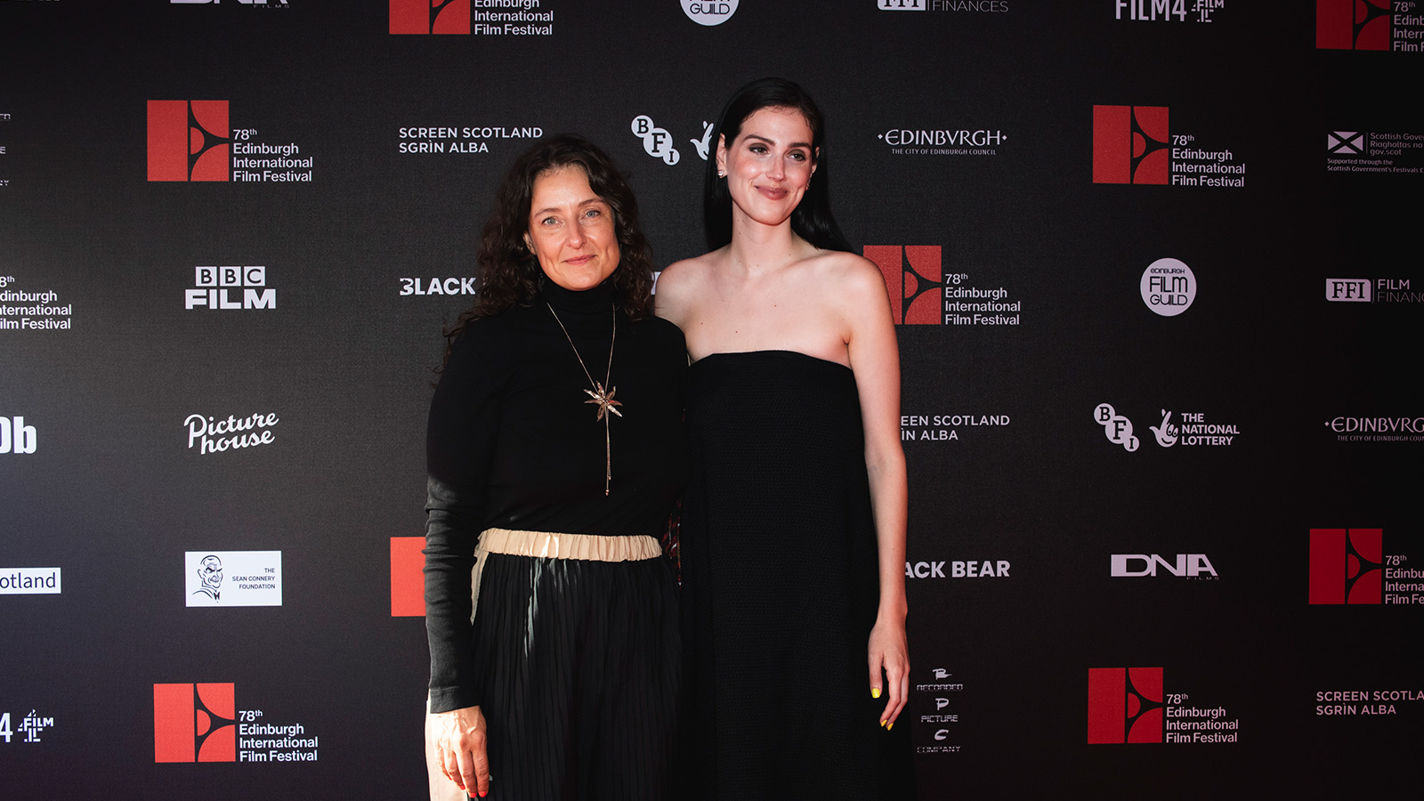 Two people posing on the red carpet in front of a backdrop with logos for the Edinburgh International Film Festival. The woman on the left wears a black turtleneck and a long necklace, while the woman on the right is in a strapless black dress.