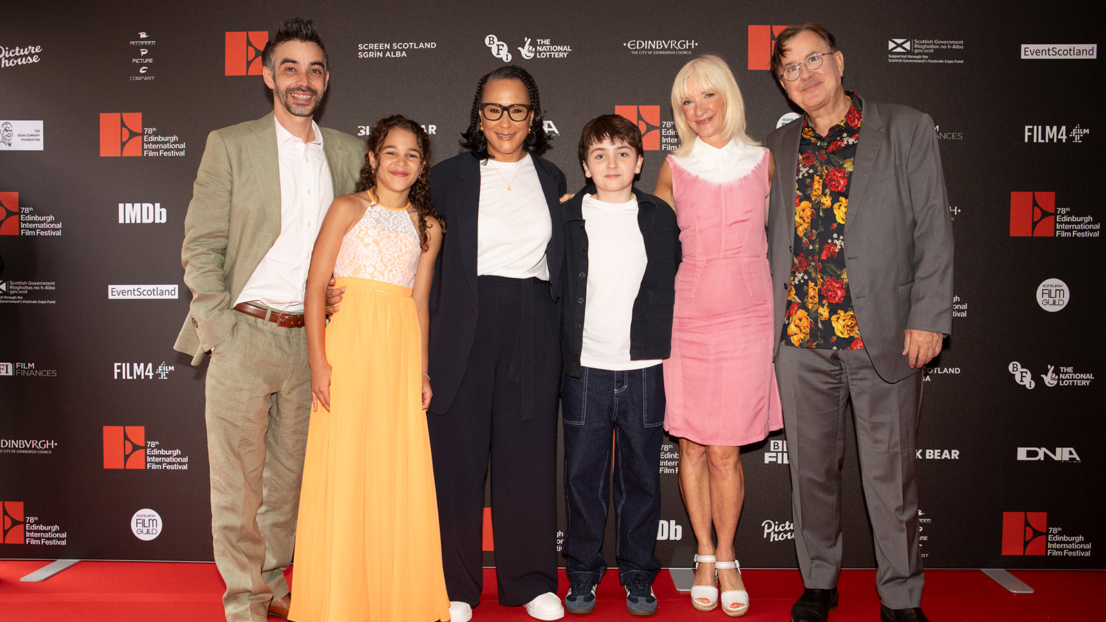 A group of six people standing together at an event. They are positioned against a black backdrop with logos, including the Edinburgh International Film Festival. The group includes a woman in a pink dress, a young boy in a dark jacket, a woman in a navy blazer, a girl in an orange dress, a man in a light suit, and another man in a grey suit with a floral shirt. They are smiling and posing for a photo.