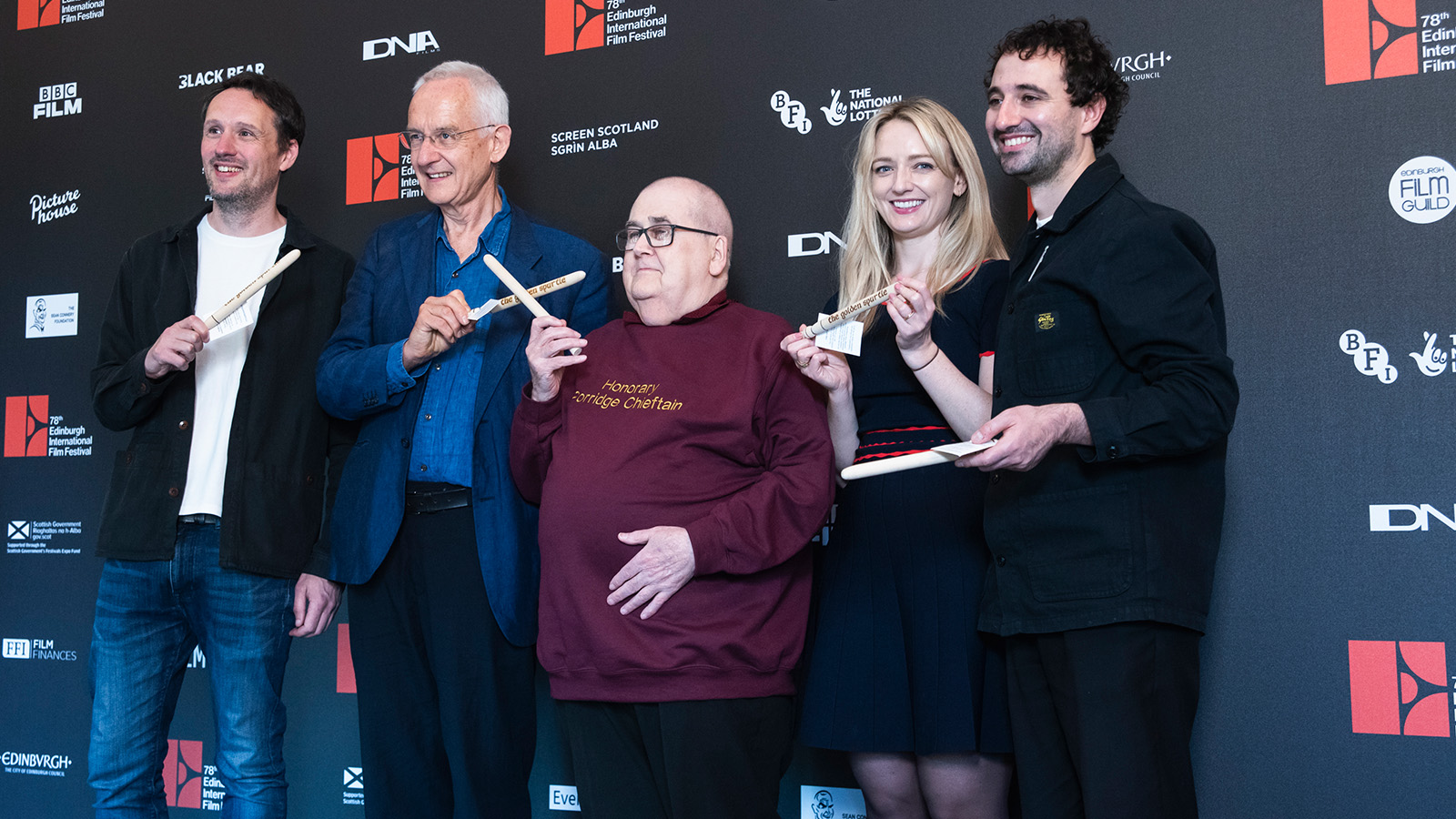 Five smiling people stand in front of a backdrop with multiple logos, holding wooden cooking utensils. The person in the center wears a sweatshirt that says "Honorary Porridge Chieftain." The backdrop includes logos for the Edinburgh International Film Festival.