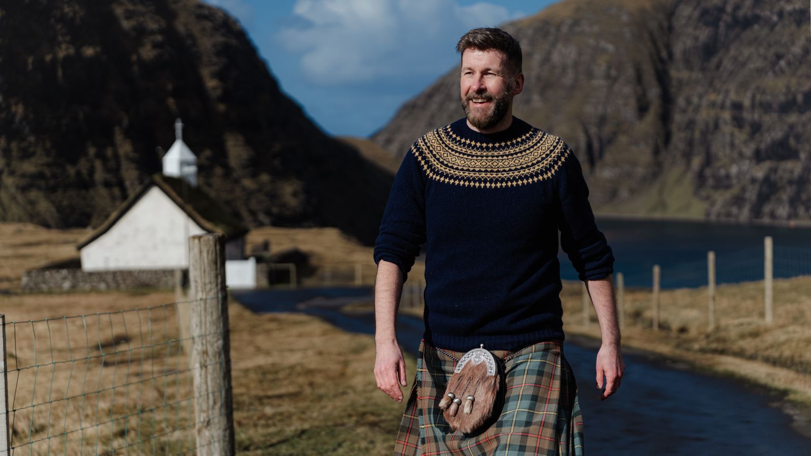 A man wearing a navy sweater with intricate yellow patterns, a kilt and sporran is walking along a rural path. He appears cheerful, with hands relaxed by his sides. In the background, there is a small white church with a pointed roof, set against a scenic landscape of hills and clear blue sky.