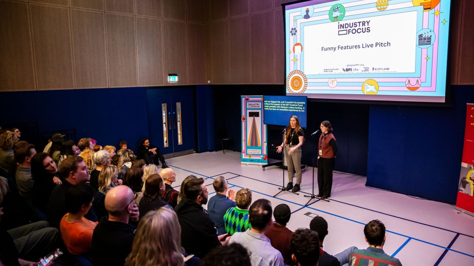 A crowd sits attentively in a small auditorium with wooden paneling. Two women stand on a small stage holding microphones, presenting during an event called 