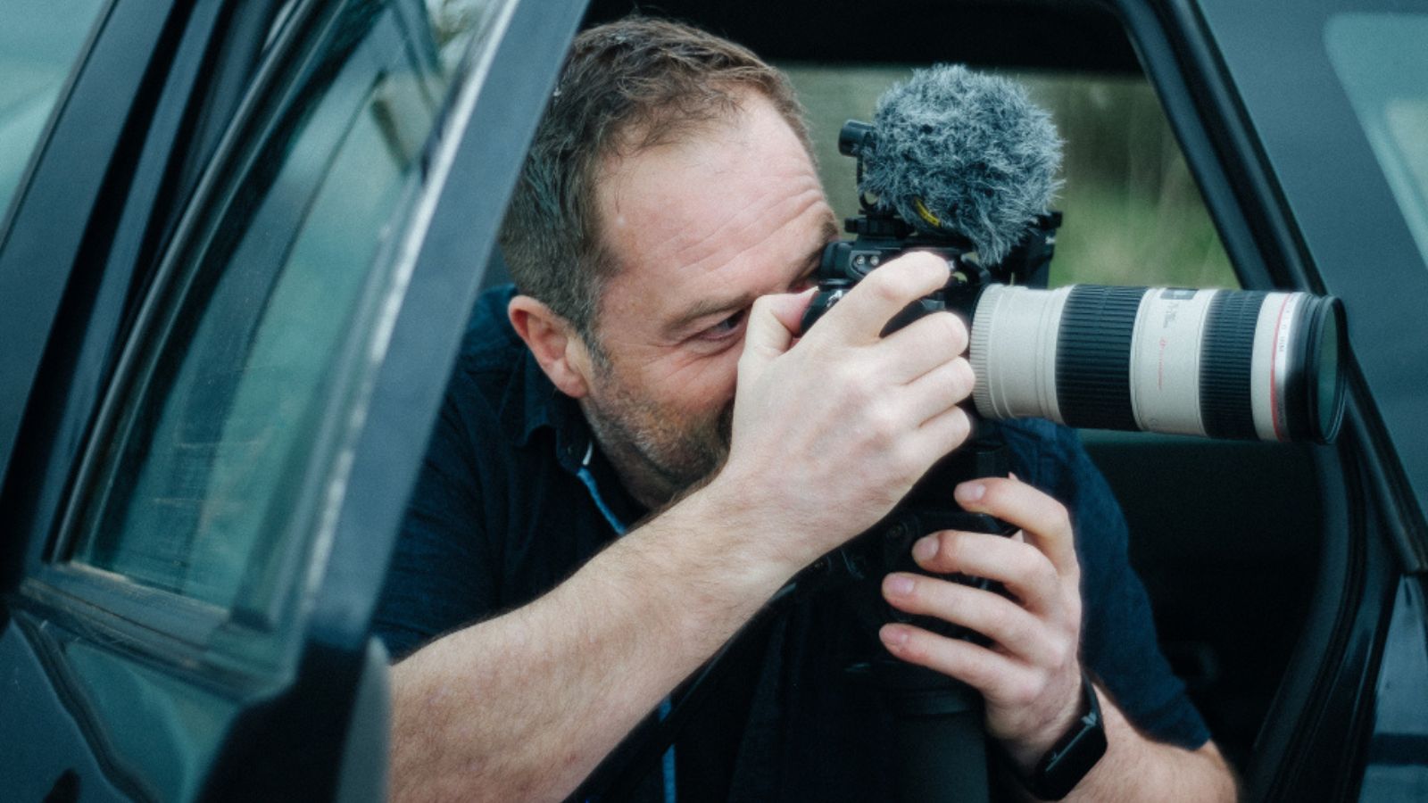 A man leans out of a car window, intently looking through a DSLR camera with a large telephoto lens. He has short brown hair and is wearing a dark shirt, focusing on capturing a shot. The camera has a furry microphone attached, suggesting video recording capabilities. The scene conveys concentration and creativity.