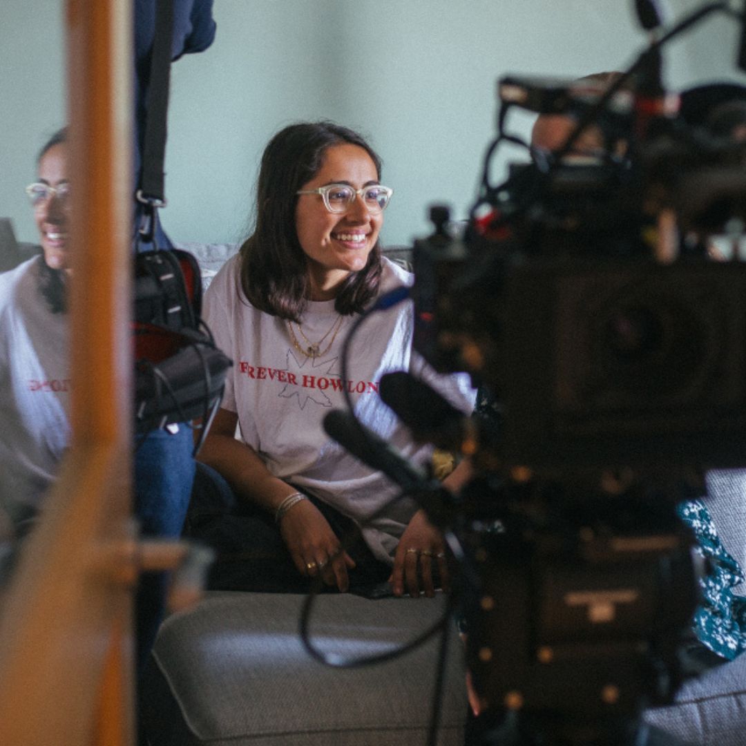 A smiling person with glasses sits on a sofa, wearing a white T-shirt with red text. They are facing a large camera setup, suggesting a filming or photography session. A reflection in the background shows the person and part of the camera equipment.