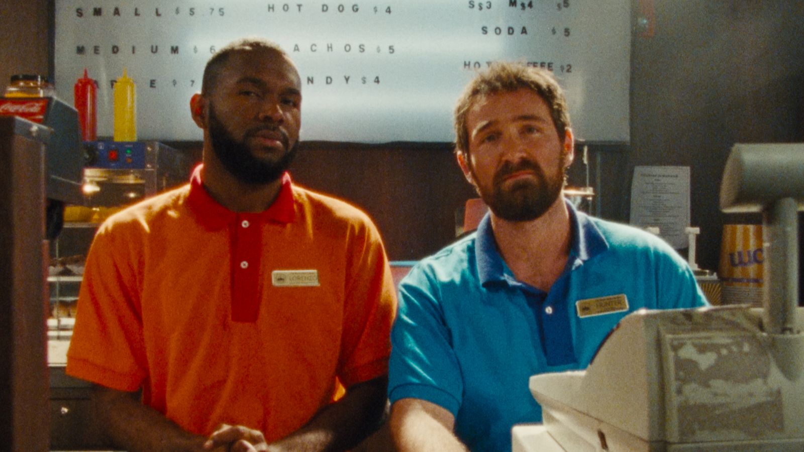 Two men stand behind a counter in a fast-food establishment. One wears an orange shirt, the other a blue shirt. A menu board reads various prices for items like hot dogs and nachos. Condiment bottles and a cash register are visible in the foreground.