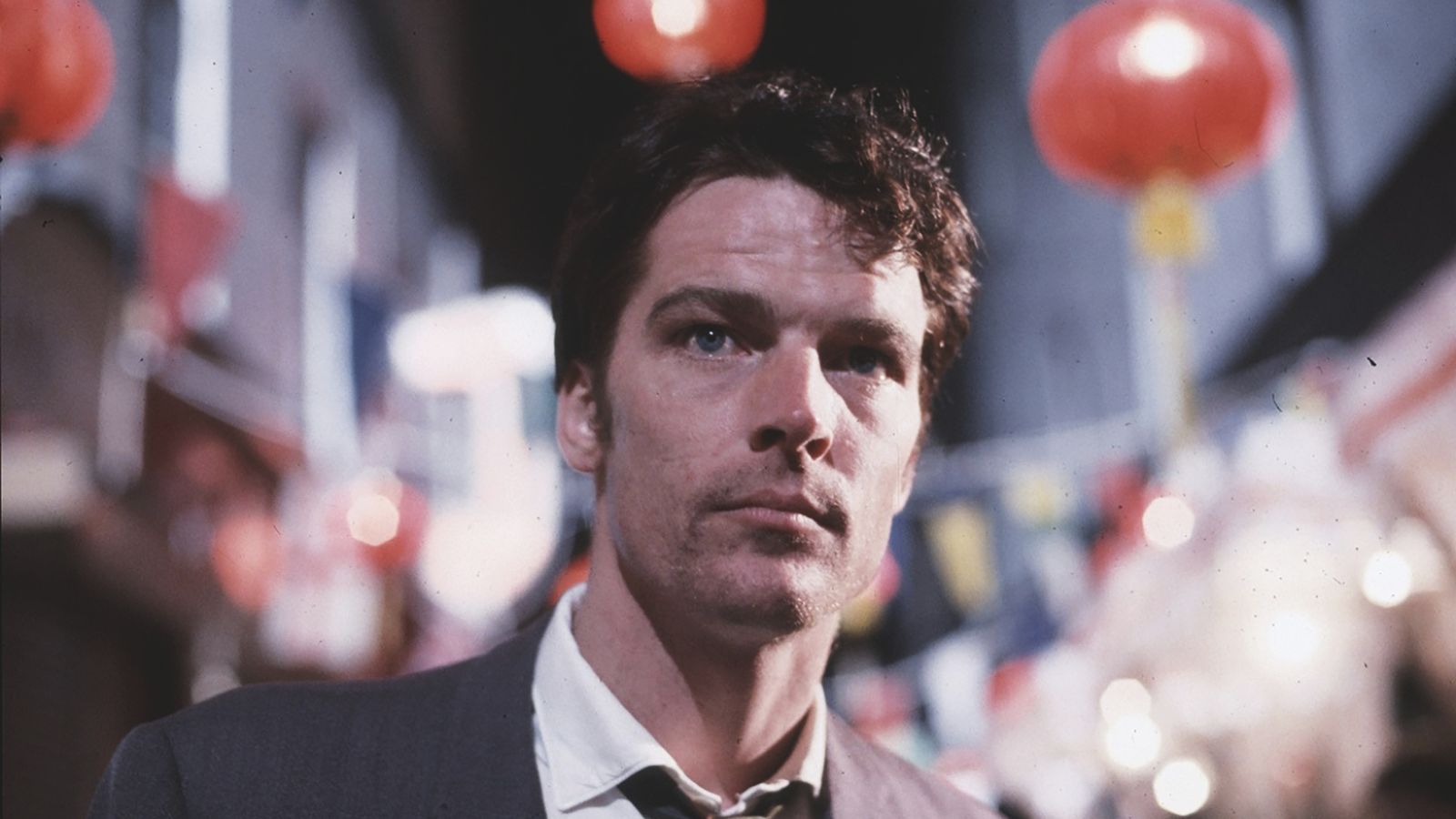 A man in a suit stands on a bustling street with red lanterns and colorful lights blurred in the background, creating a festive atmosphere.