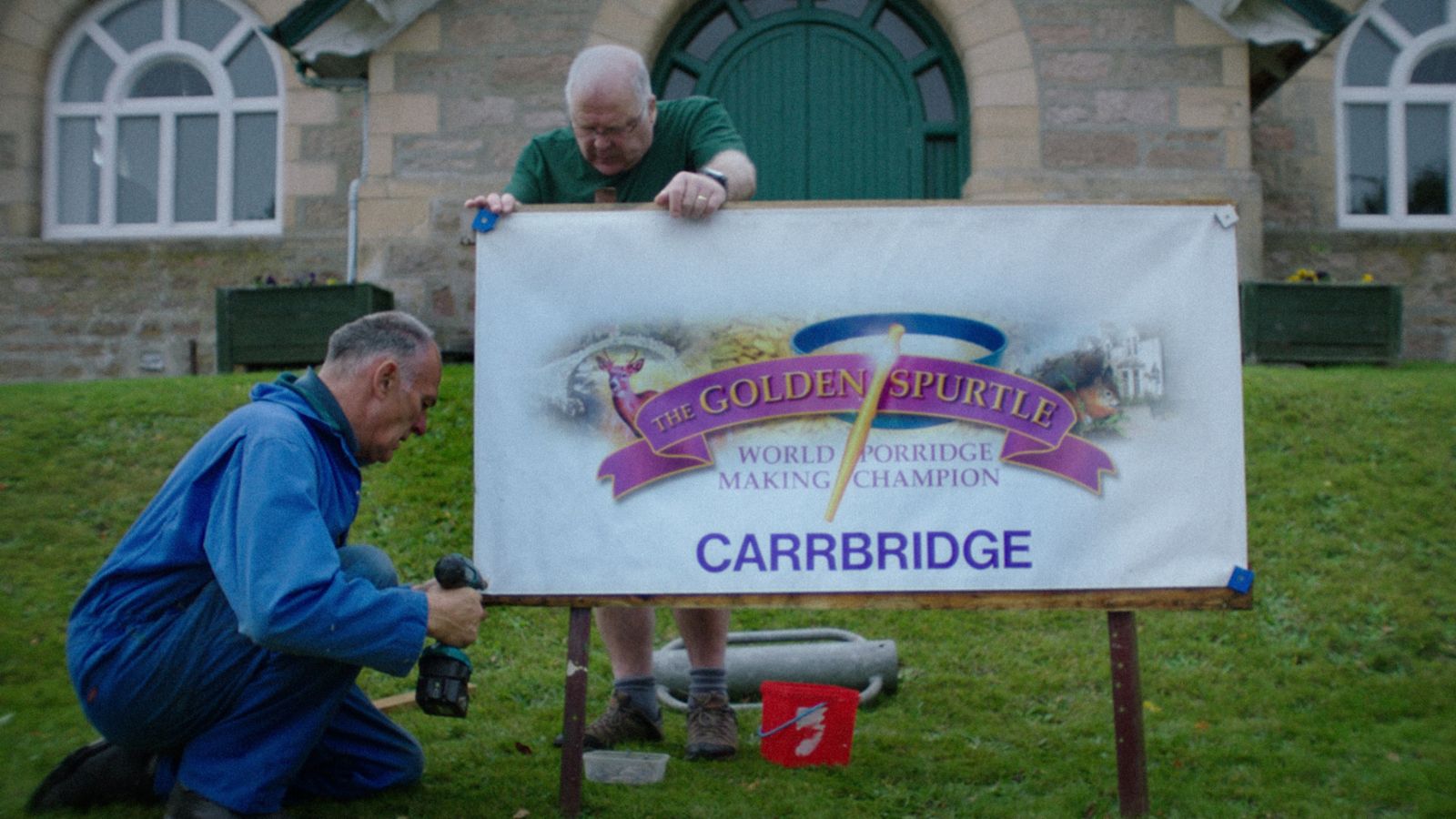 Two men set up a sign for "The Golden Spurtle: World Porridge Making Champion" in Carrbridge. One man holds the sign while the other, dressed in blue coveralls, kneels with a power tool. They are in front of a stone building with arched windows and a green door.