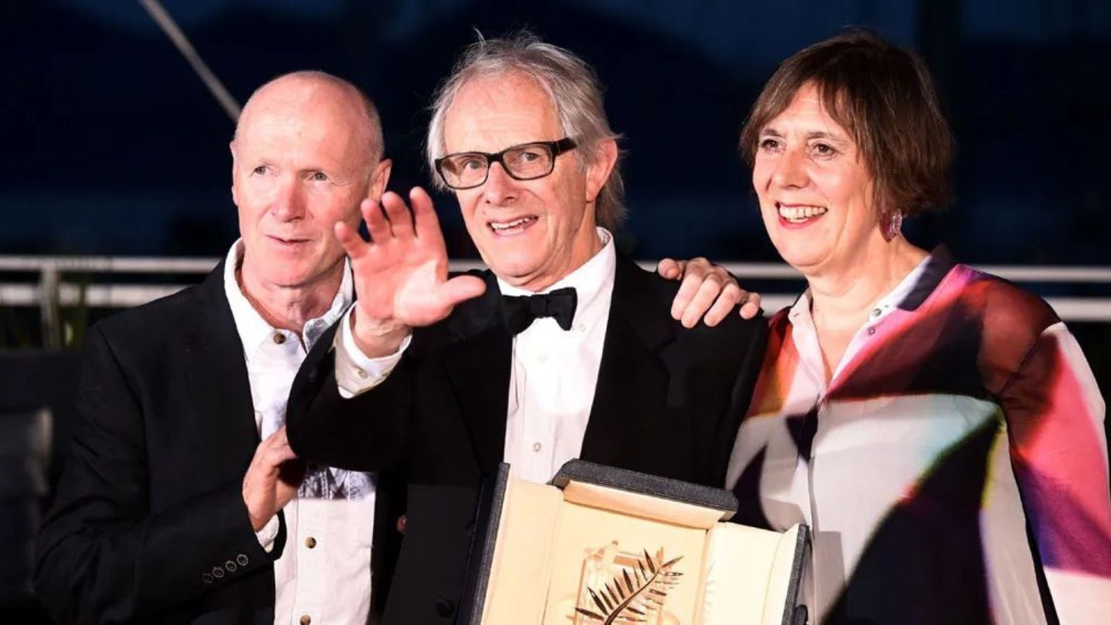 Paul Laverty, Ken Loach and Rebecca O’Brien stand in a row. Paul and Ken wear black suits with white shirts, while Rebecca wears a red and blue blouse.