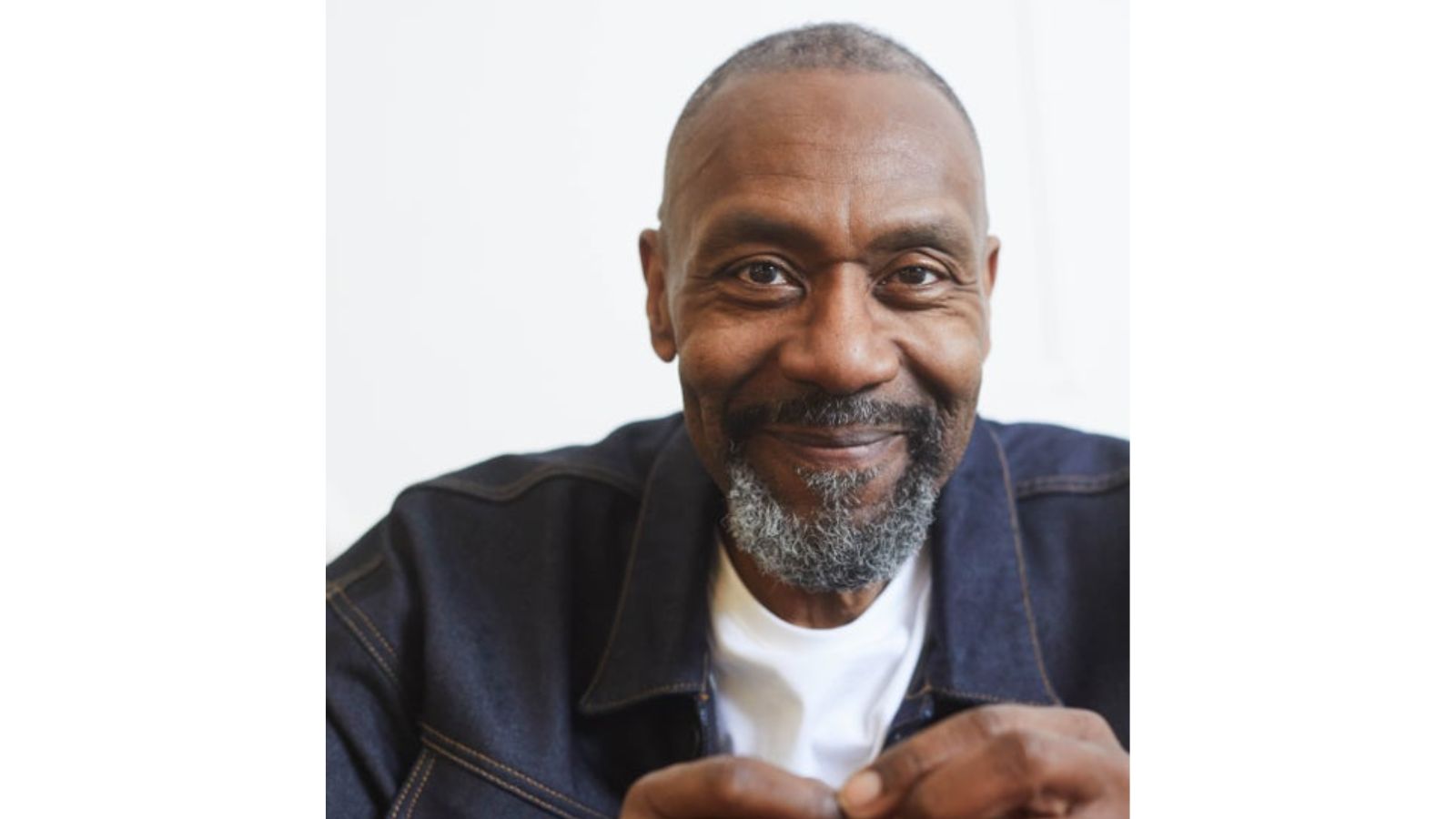 A smiling man with a beard and short hair is wearing a dark denim jacket over a white shirt. He looks directly at the camera, with his hands visible in the foreground. The background is plain white.