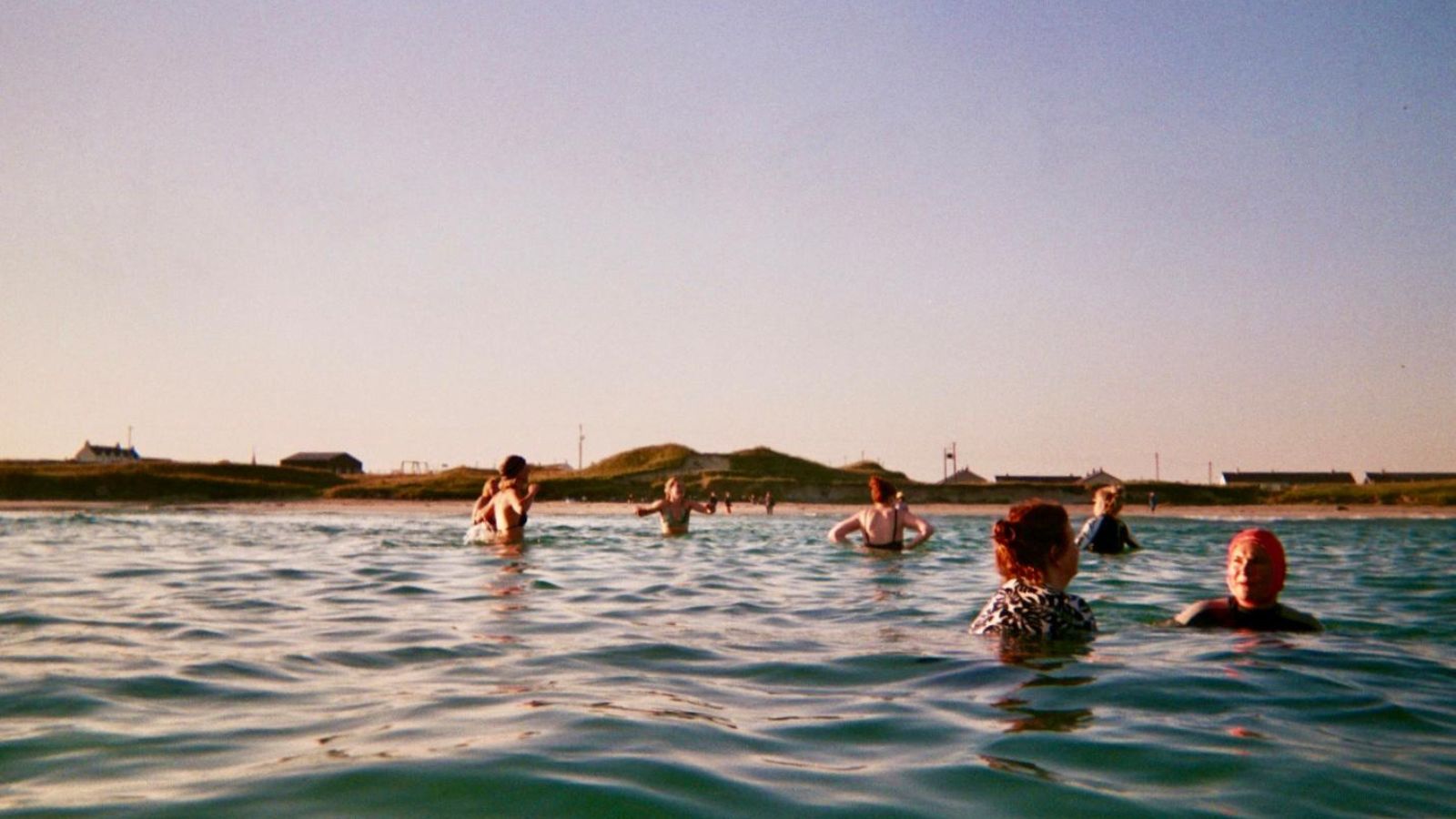 A group of people swims in the ocean near the coast, with grassy dunes and buildings visible in the background under a clear blue sky. The water is calm, and the setting suggests a sunny day.
