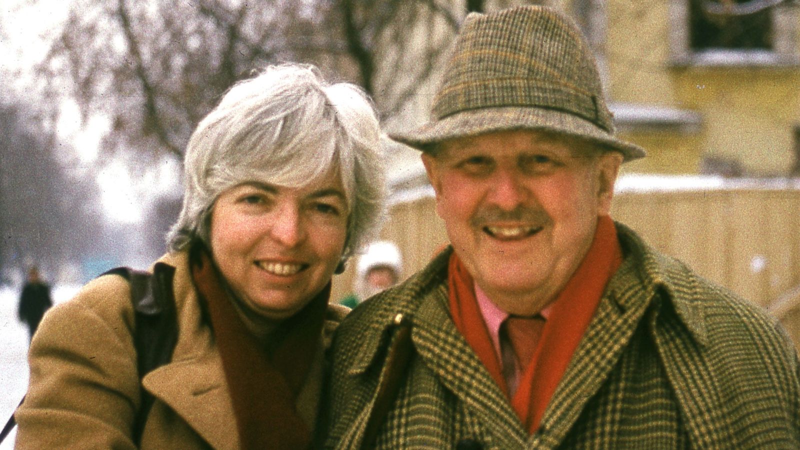 A smiling older couple stands outdoors in winter. The woman has short gray hair and wears a tan coat with a scarf, while the man wears a plaid coat and hat. Snowy landscape and blurred trees are visible in the background.