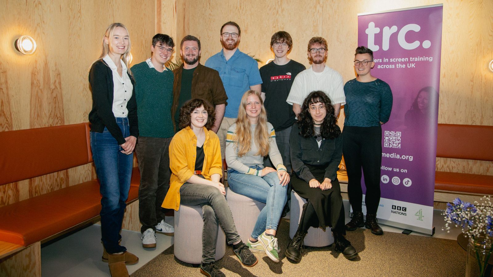 A group of ten people are posing in a warmly lit room with wooden walls and an orange bench. Four are seated on light-colored stools, and six are standing behind them. They are all smiling and appear casual. A purple banner on the right reads 