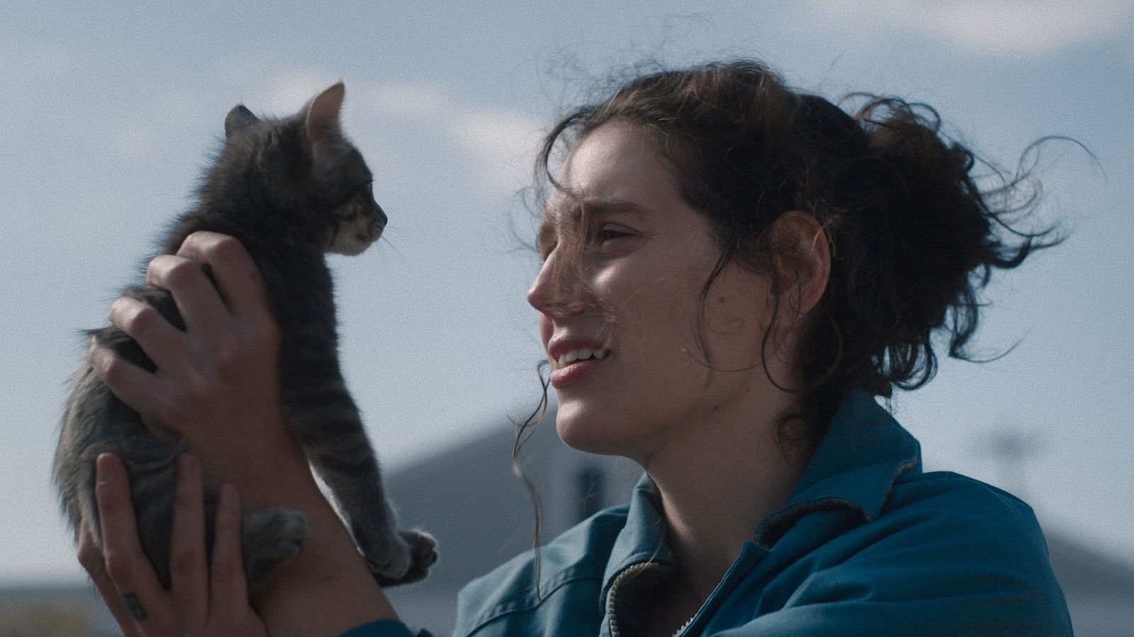 A person holds a small tabby kitten close, both gazing at each other, set against a blurred outdoor background.