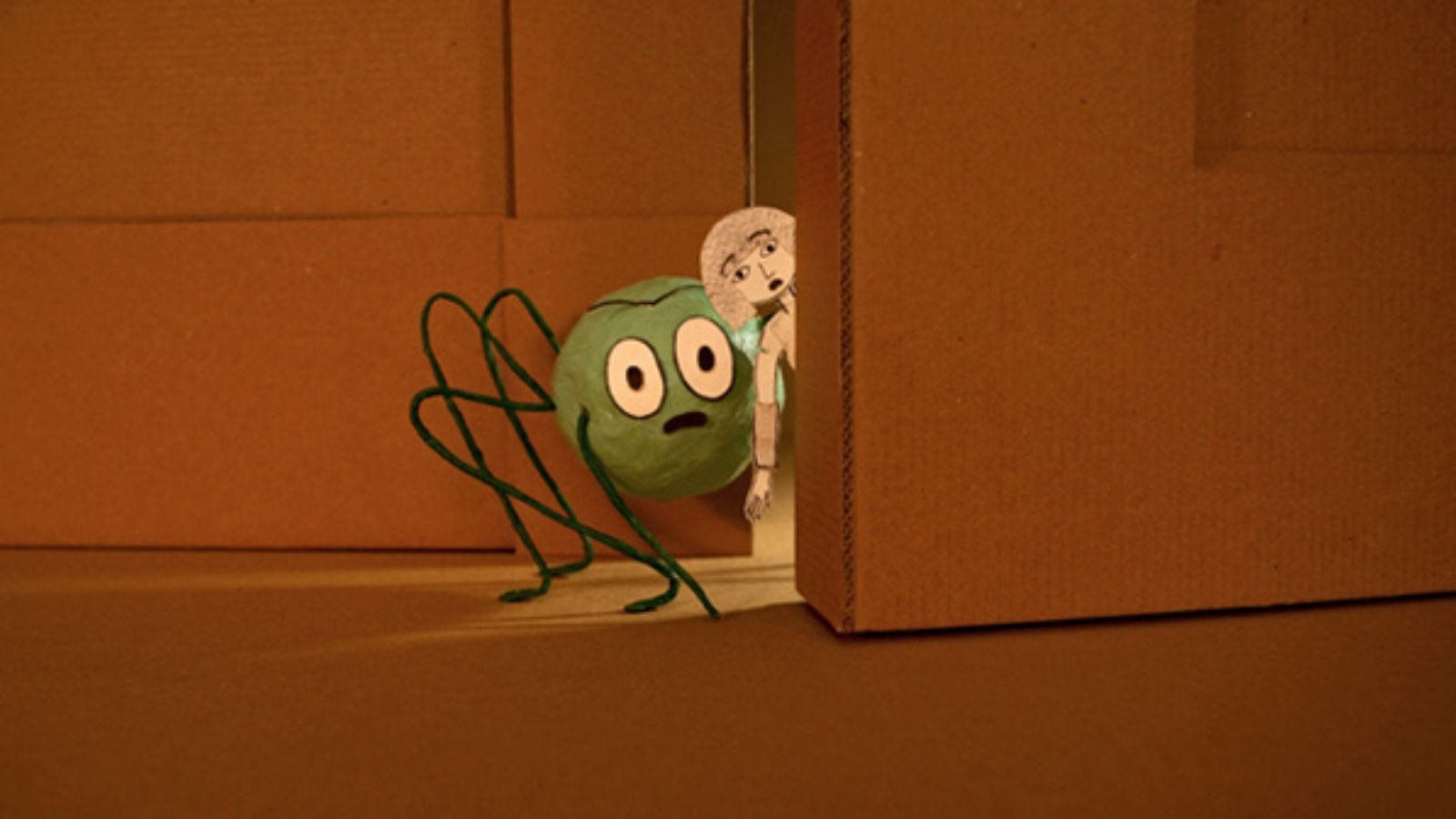 A whimsical, green, round character with large eyes and thin wire limbs appears peering out from behind a slightly opened cardboard door. Beside it, a small, monochrome cutout figure with a surprised expression also peeks out. The background is composed of cardboard, creating a simple, crafty scene.
