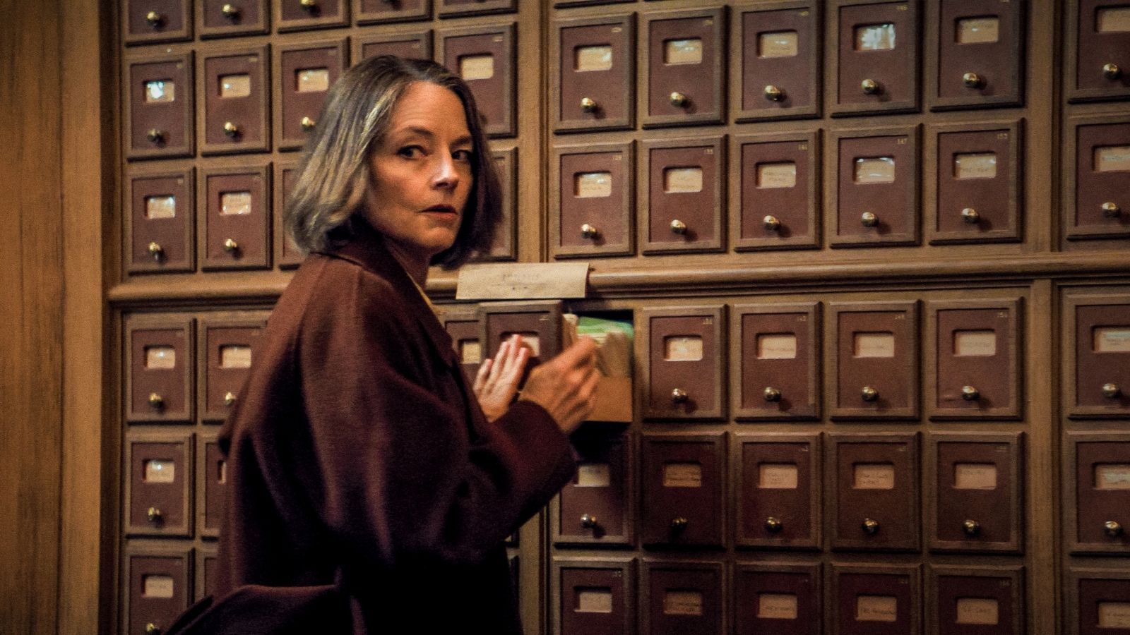 A woman stands in front of a large wall of vintage wooden mailboxes, each with a small brass knob. She holds a green envelope and looks over her shoulder, conveying a sense of urgency or curiosity.