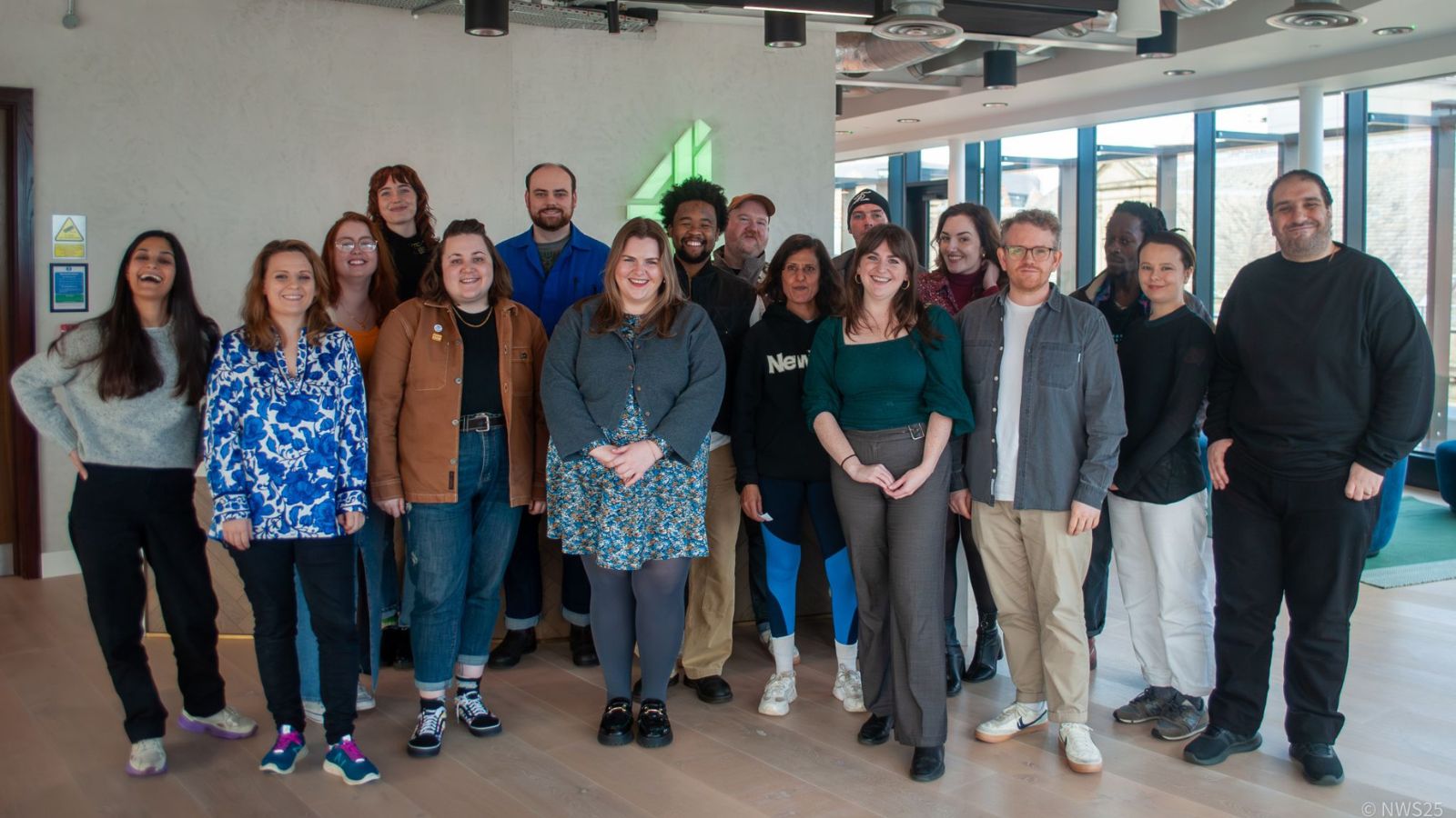 A diverse group of eighteen people poses indoors in a bright room with large windows. They appear cheerful and relaxed, standing closely together. The setting includes modern furnishings and a stylised green number four on the wall behind them. The group is casually dressed, showcasing a variety of clothing styles and colours.