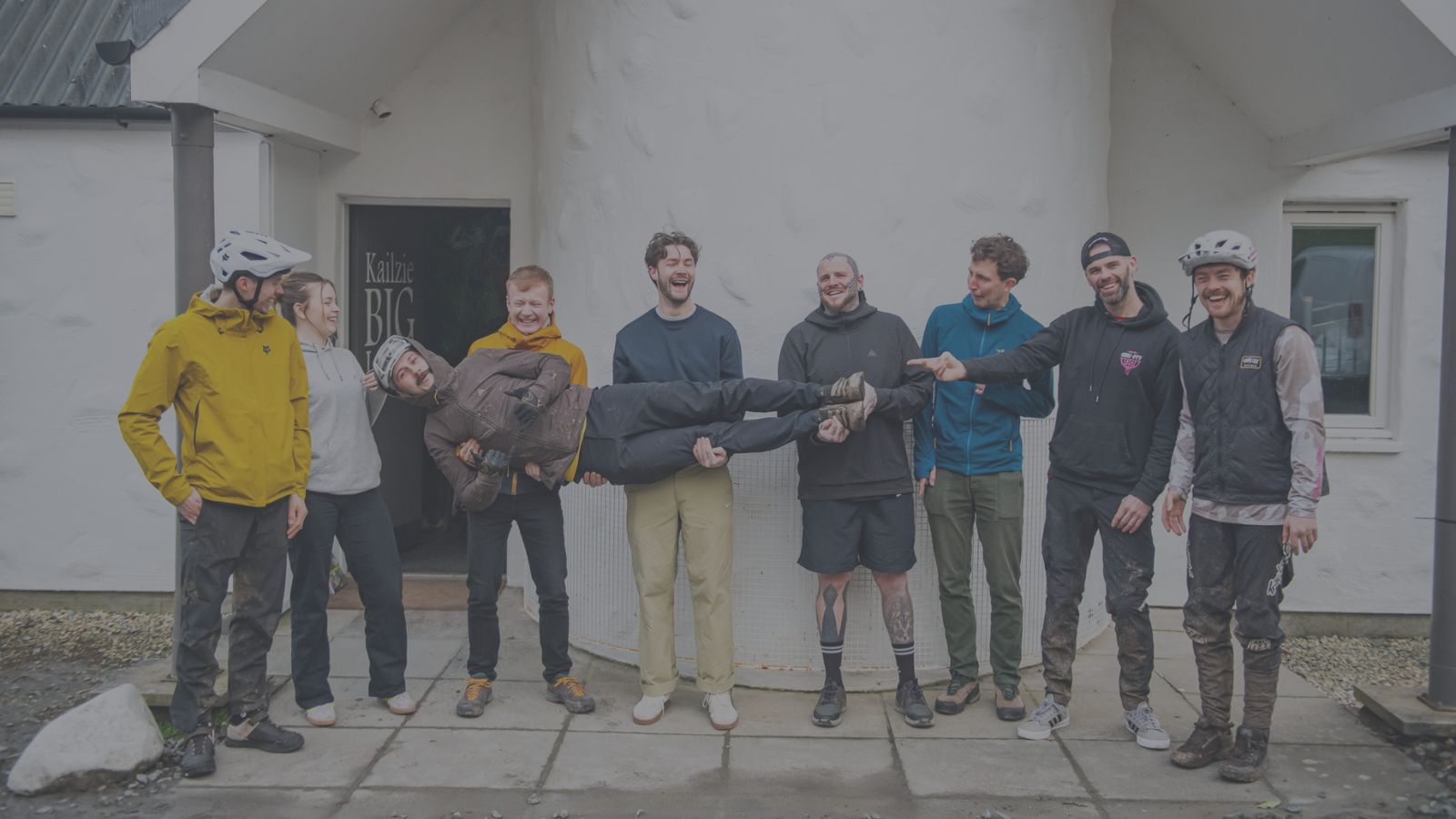 A group of eight people stand outside a white building. Seven are on their feet, while one person is held horizontally by four others at the centre. Some are wearing helmets and outdoor gear, and everyone appears to be laughing or smiling.