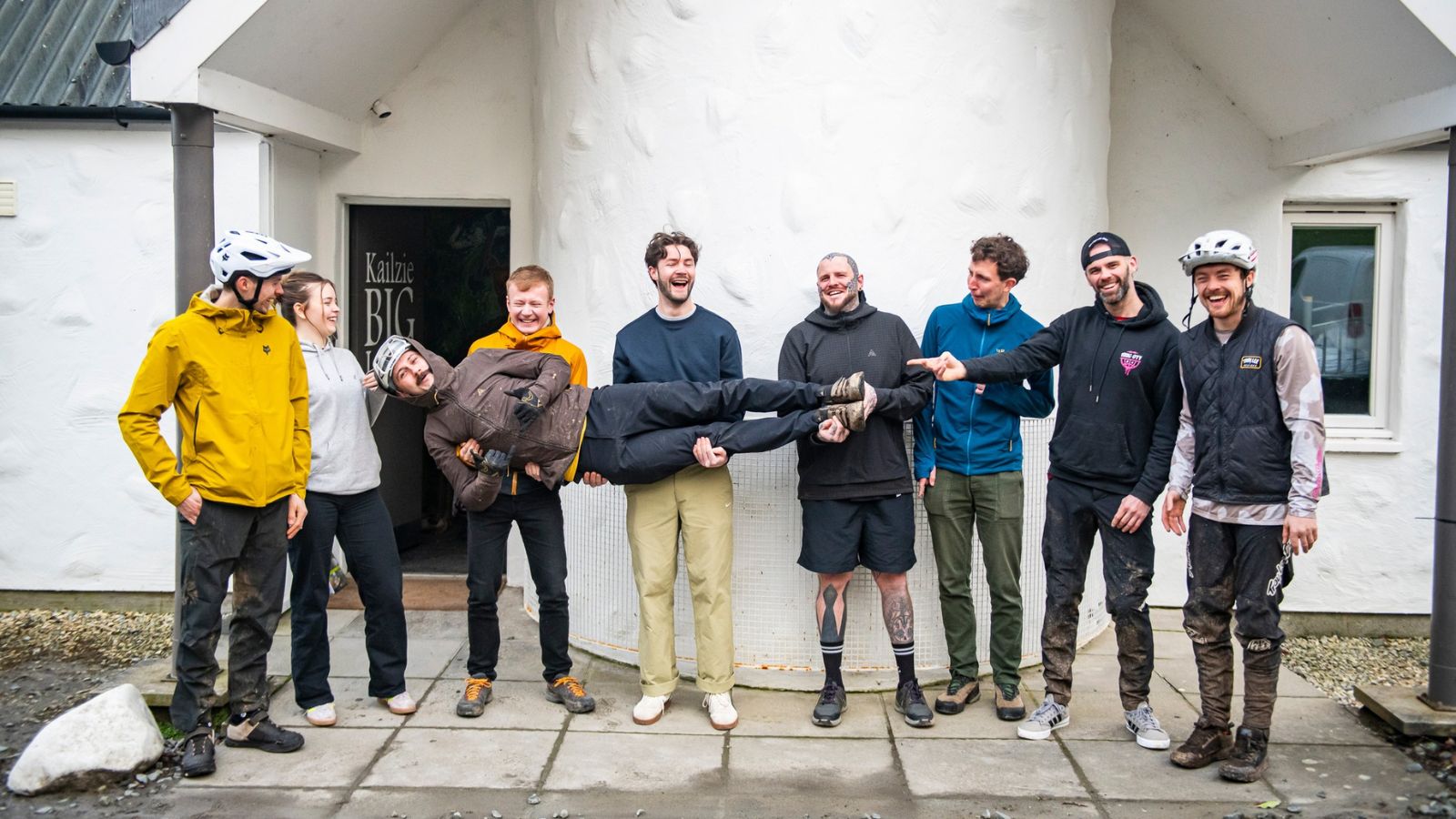 A group of eight people stand outside a white building. Seven are on their feet, while one person is held horizontally by four others at the centre. Some are wearing helmets and outdoor gear, and everyone appears to be laughing or smiling.