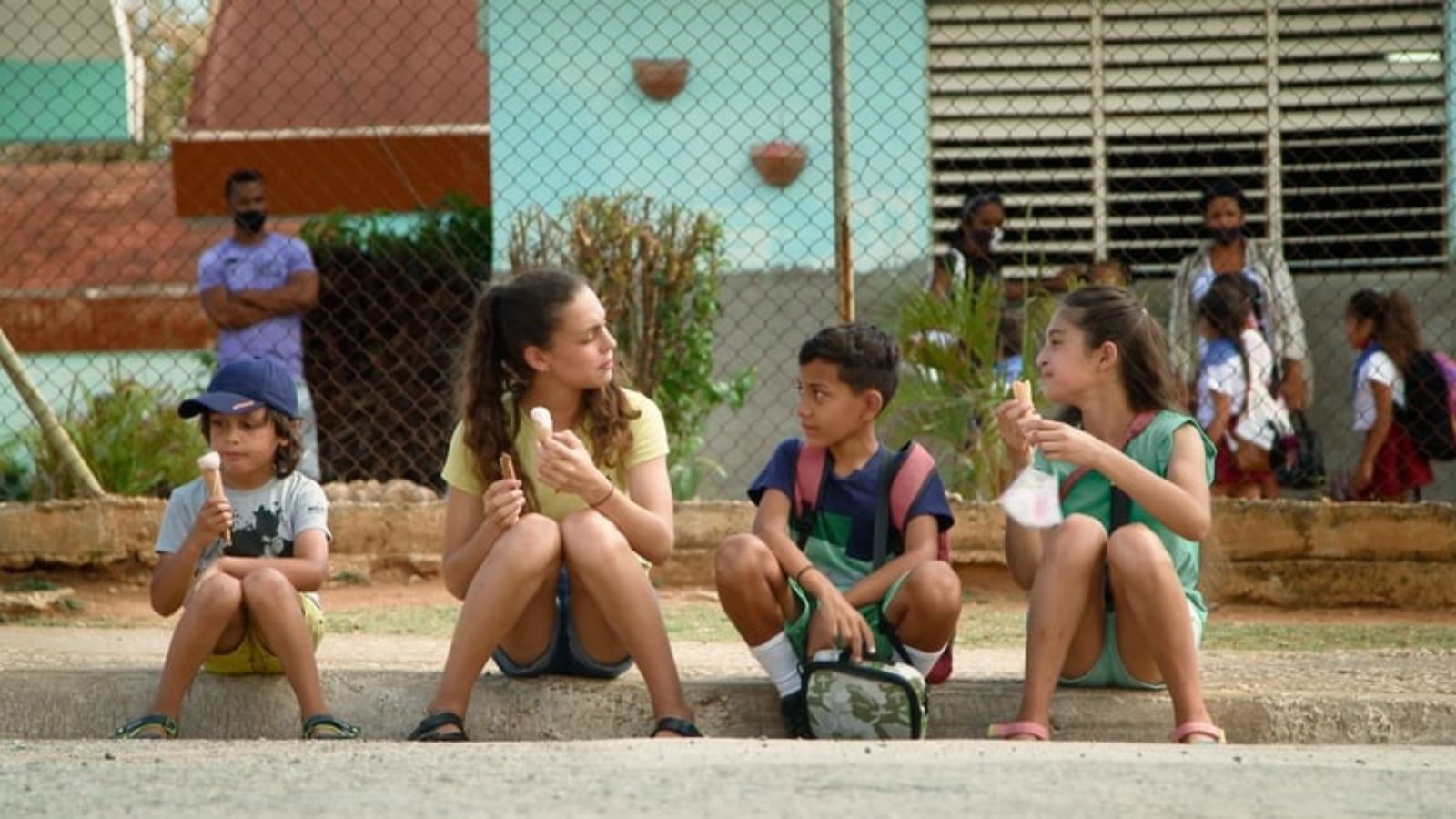 our children sit on a curb, each holding an ice cream cone. They are casually dressed and appear relaxed. In the background, a chain-link fence separates them from a group of people, including some children in school uniforms and an adult in a purple shirt. The scene takes place outdoors, with a light blue building and plants behind the fence.