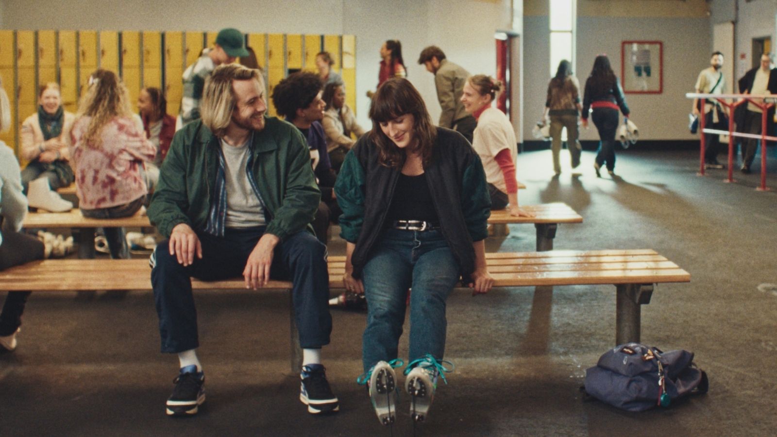 A lively ice rink locker room with people sitting on wooden benches. In the foreground, a man and woman share a smile. The woman is wearing ice skates, their teal laces visible. Behind them, others chat against a backdrop of yellow lockers. A sense of camaraderie fills the space.