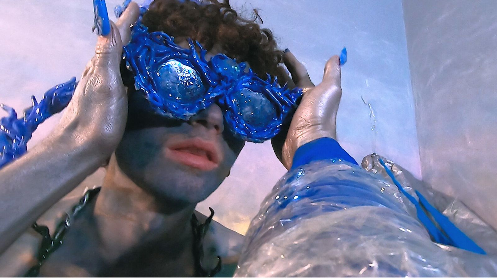 A person with curly hair wears large, intricate blue goggles. Their hands, painted silver, adjust the goggles. The blue and silver colours are striking against a muted background, creating an artistic and unusual visual effect.