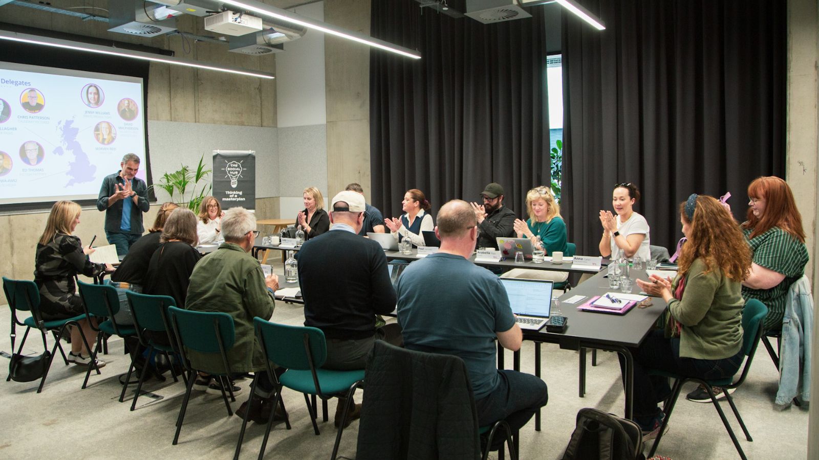 A group of people seated around a rectangular conference table in a modern meeting room, some clapping and others focused on laptops. A presenter stands in front of a screen displaying a slide with headshots and a map. The room has industrial-style decor with concrete walls and dark curtains.