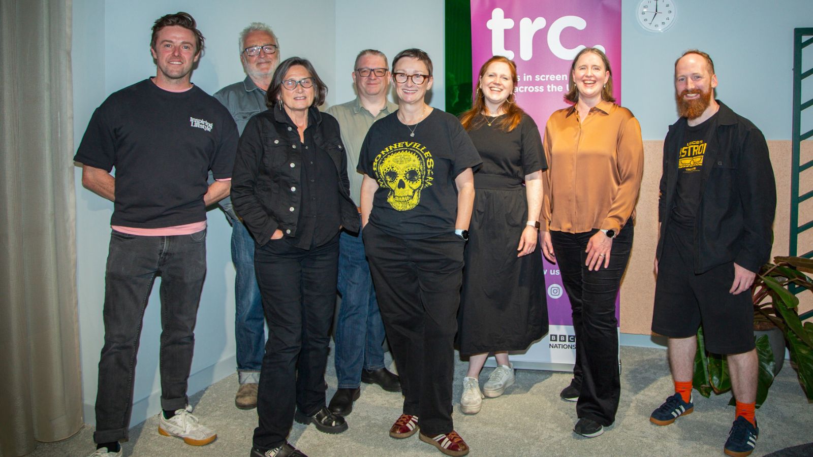 Eight people stand together in a casual group, smiling for the camera. They are in front of a banner with the letters 