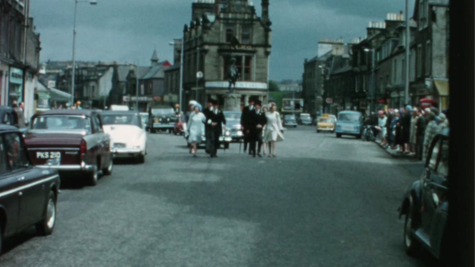 A vintage street scene depicts a parade with people walking down the middle of a wide road lined with parked classic cars. Onlookers stand on both sidewalks, observing the activity. The sky is overcast, adding a dramatic backdrop to the historic architecture in the background.