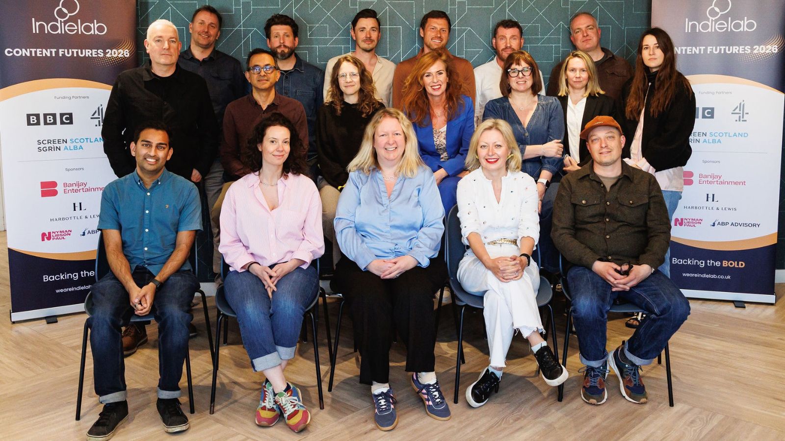 A group of eighteen people are posed together, most sitting, some standing, smiling towards the camera. They are in casual attire, with a background featuring two vertical banners reading “Indielab Content Futures 2026” and logos of sponsors like BBC and Channel 4. The setting suggests a collaborative, professional environment.