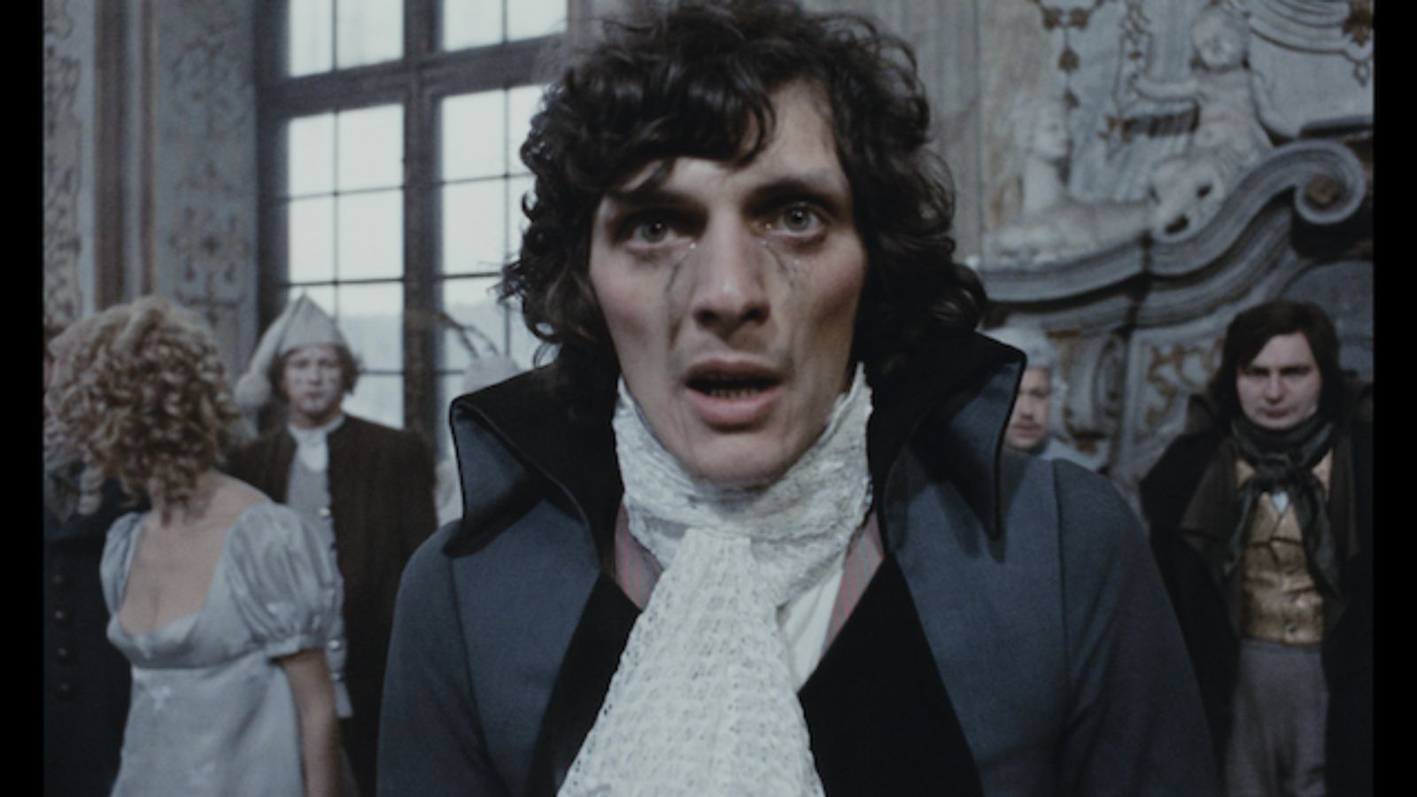 A man with curly hair and a distressed expression, wearing a high-collared coat and a white cravat, stands indoors. Behind him are people in period attire, including a woman in a light dress and a man in a hat, amidst an ornate setting with large windows and detailed carvings.