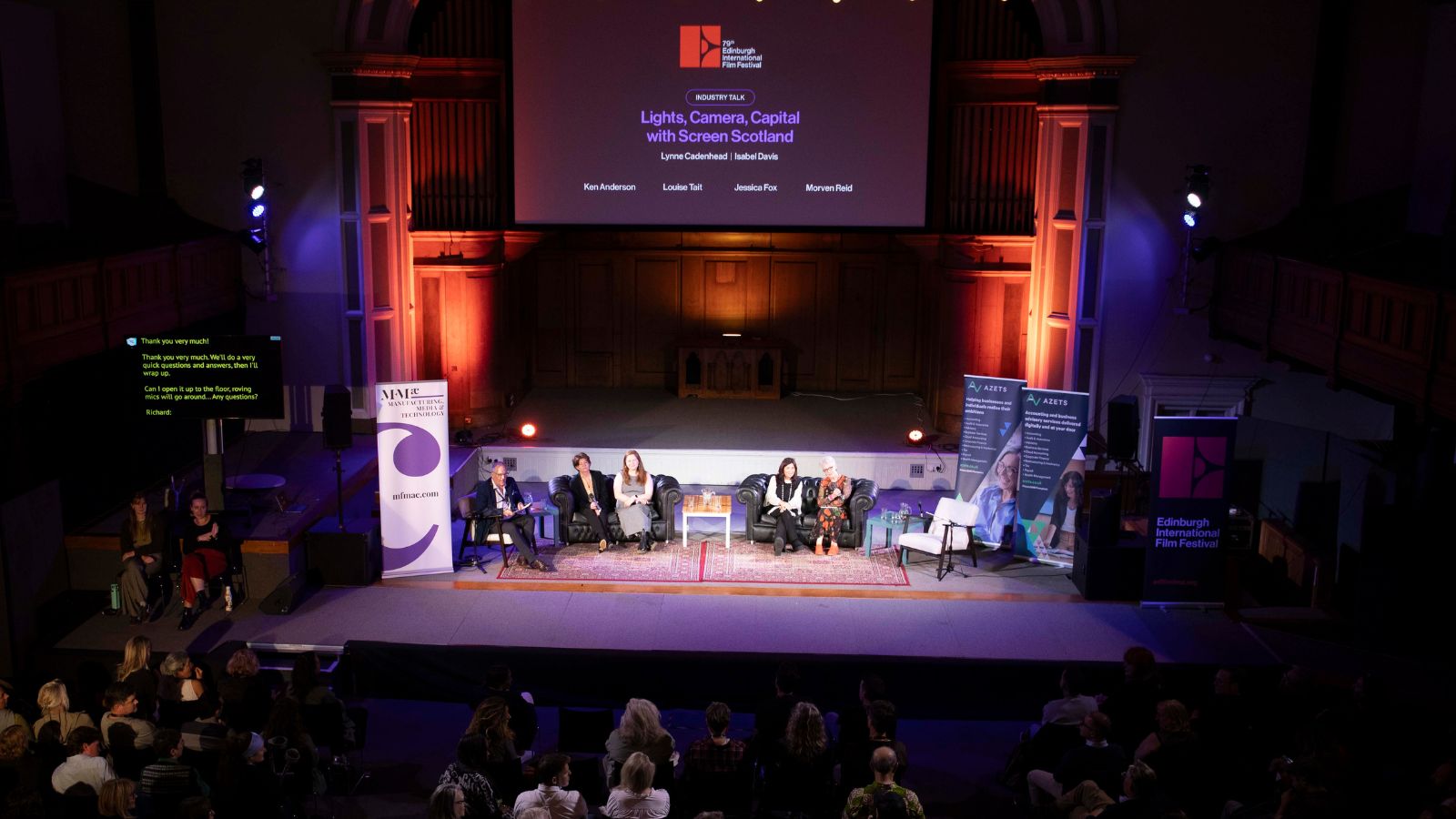 An elevated, wide-angle shot of a panel discussion at the 79th Edinburgh International Film Festival, held on a stage inside a dimly lit auditorium with orange and blue spotlighting. A large projection screen above the stage reads, "INDUSTRY TALK: Lights, Camera, Capital with Screen Scotland," featuring names Lynne Cadenhead, Isabel Davis, Ken Anderson, Louise Tait, Jessica Fox, and Morven Reid. On the stage, five panelists sit in dark armchairs on a patterned red rug, with small wooden side tables between them. Banners for Azets and MBM Commercial frame the stage on both sides. In the foreground, the backs of the audience members are visible as they sit in the dark, watching the presentation.