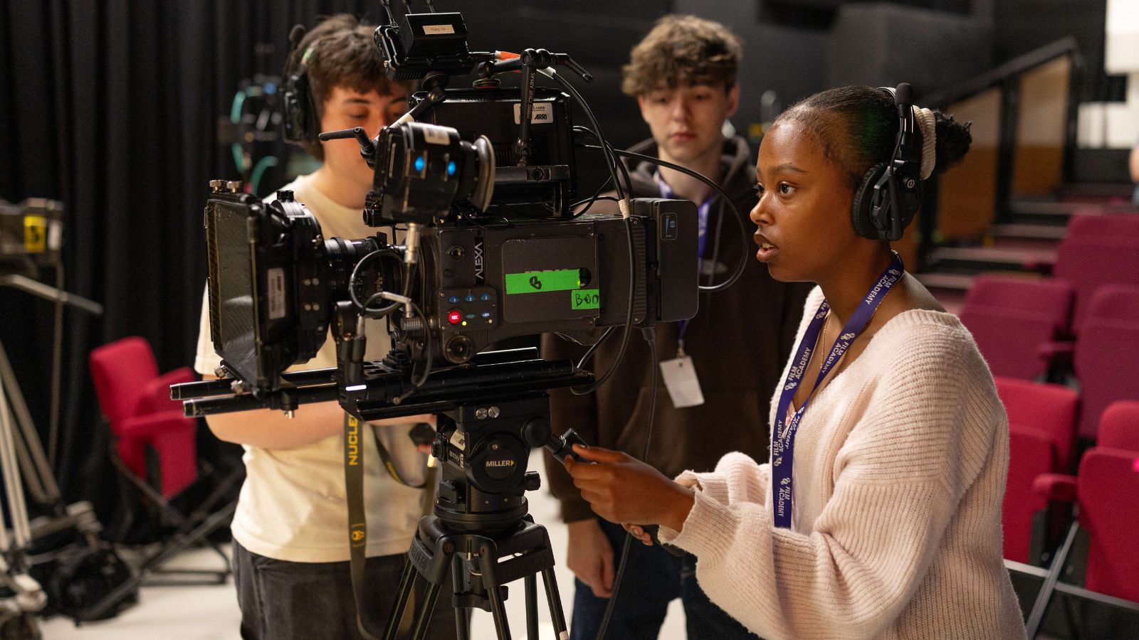 Medium shot of three young film students operating a large professional ARRI Alexa camera on a Miller tripod in an indoor space.