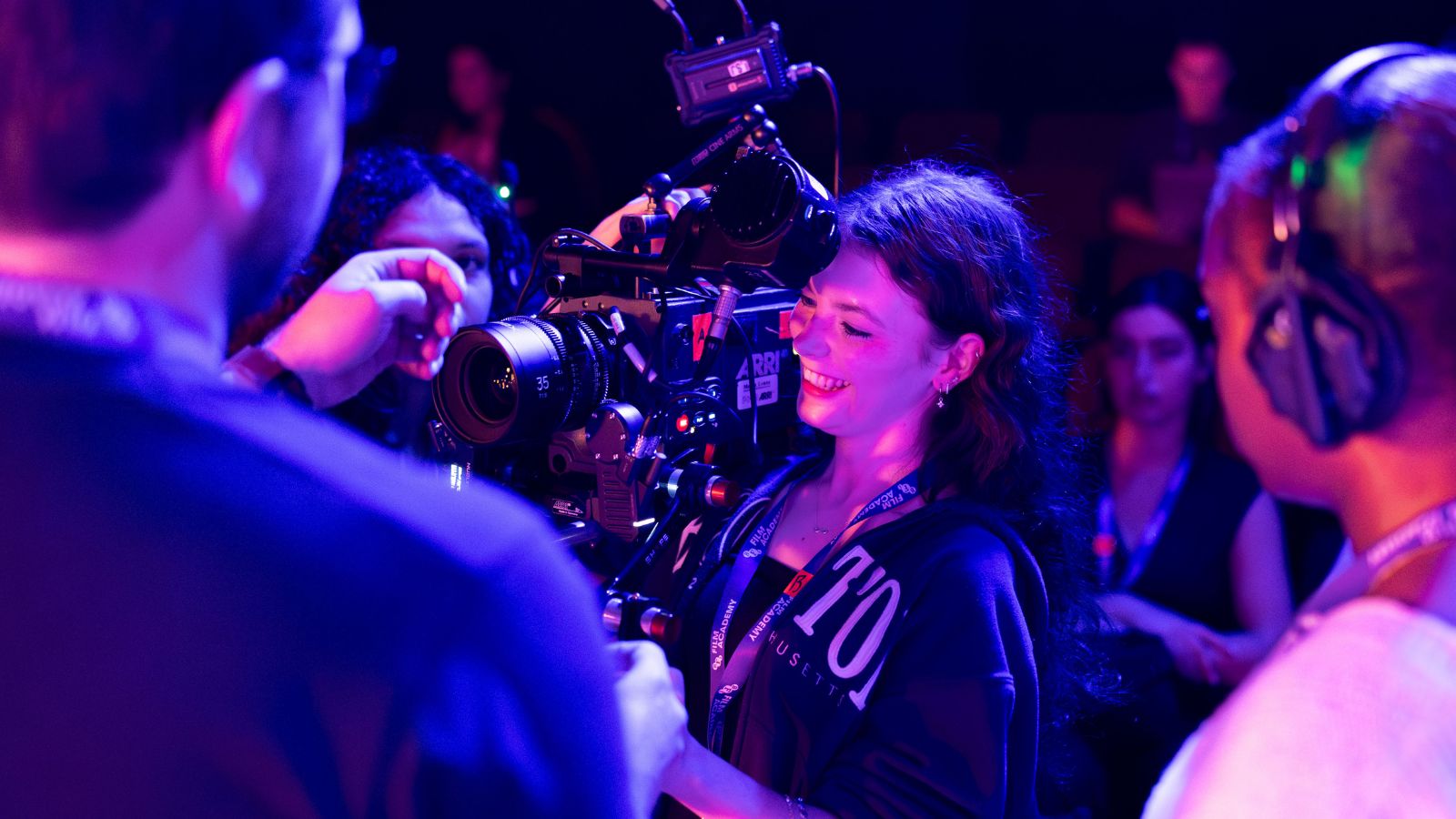 An over-the-shoulder medium shot under vibrant purple and blue stage lighting shows a smiling young woman with long, wavy dark hair operating a large ARRI professional cinema camera. Other crew members are gathered around her in a dimly lit auditorium, with some hands adjusting the camera's 35mm lens.