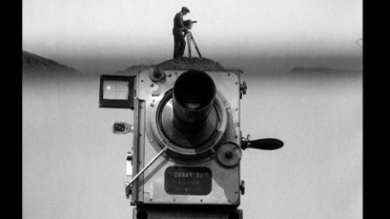 A black-and-white double-exposure image featuring a large, vintage movie camera in the foreground. Atop the camera housing stands a tiny silhouette of a cameraman operating another camera on a tripod, appearing as if he is standing on a hill against a hazy, distant horizon.