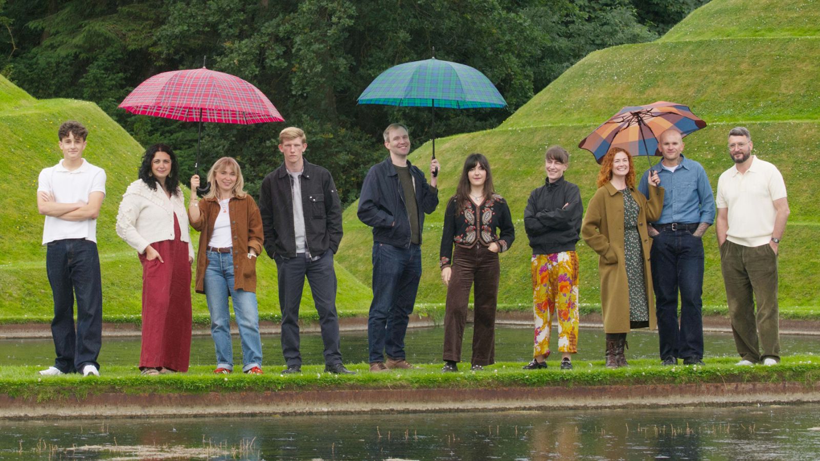 An eye-level shot captures ten people standing in a row in an outdoor setting with rolling green grassy hills and a small body of water in the foreground, under an overcast sky.